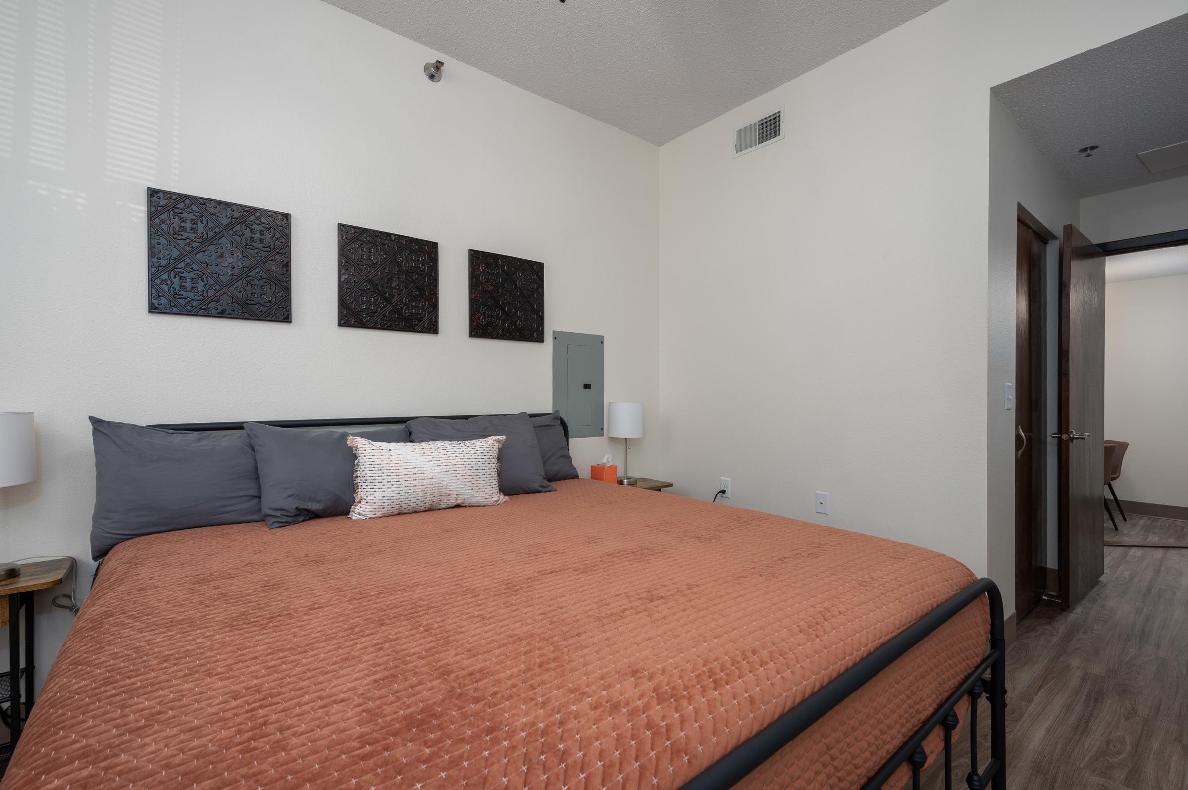 A cozy bedroom featuring a large bed with an orange quilt, gray pillows, and a decorative throw pillow. Three framed artworks are mounted on the pale wall above the bed. Two bedside lamps illuminate the space. A doorway leads to another room, suggesting a larger living area.