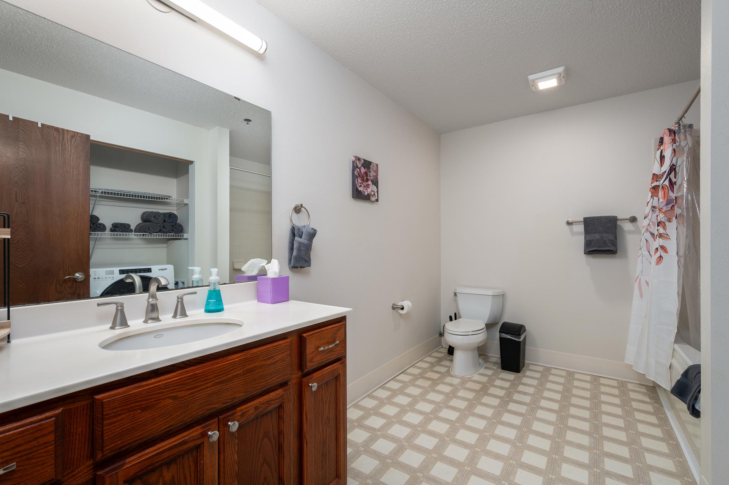A spacious bathroom featuring a white sink with a wooden vanity, a large mirror, and a gray shower curtain. There are neatly stacked towels on shelves, a toilet, and a small trash can. The light-colored walls and tiled floor create a clean, modern atmosphere.