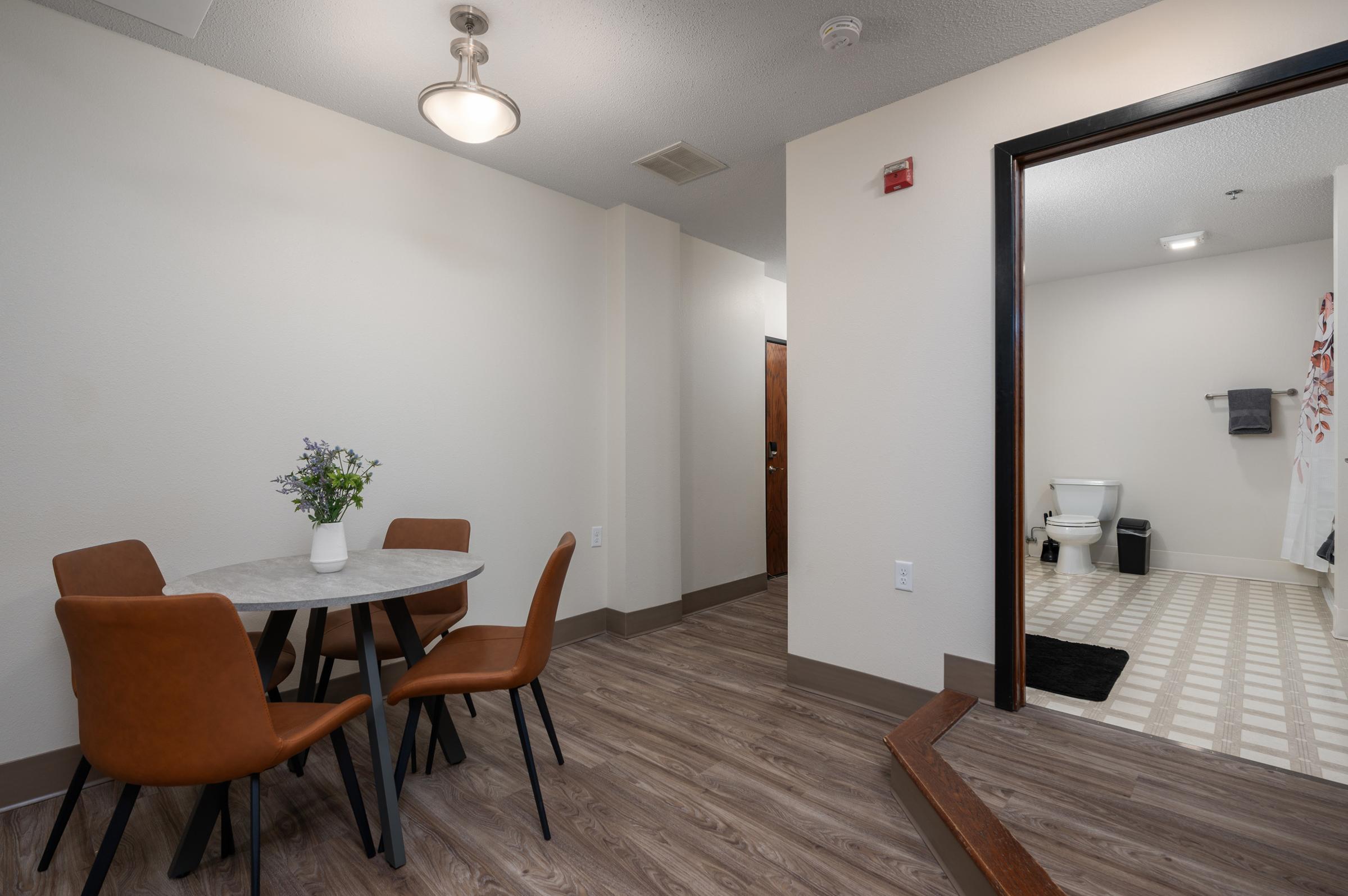 A small dining area featuring a round marble table with four brown chairs. In the background, a hallway leads to a bathroom, which is visible through an open door. The bathroom includes a toilet and is styled with light-colored tiles. Natural light illuminates the space, creating a warm atmosphere.