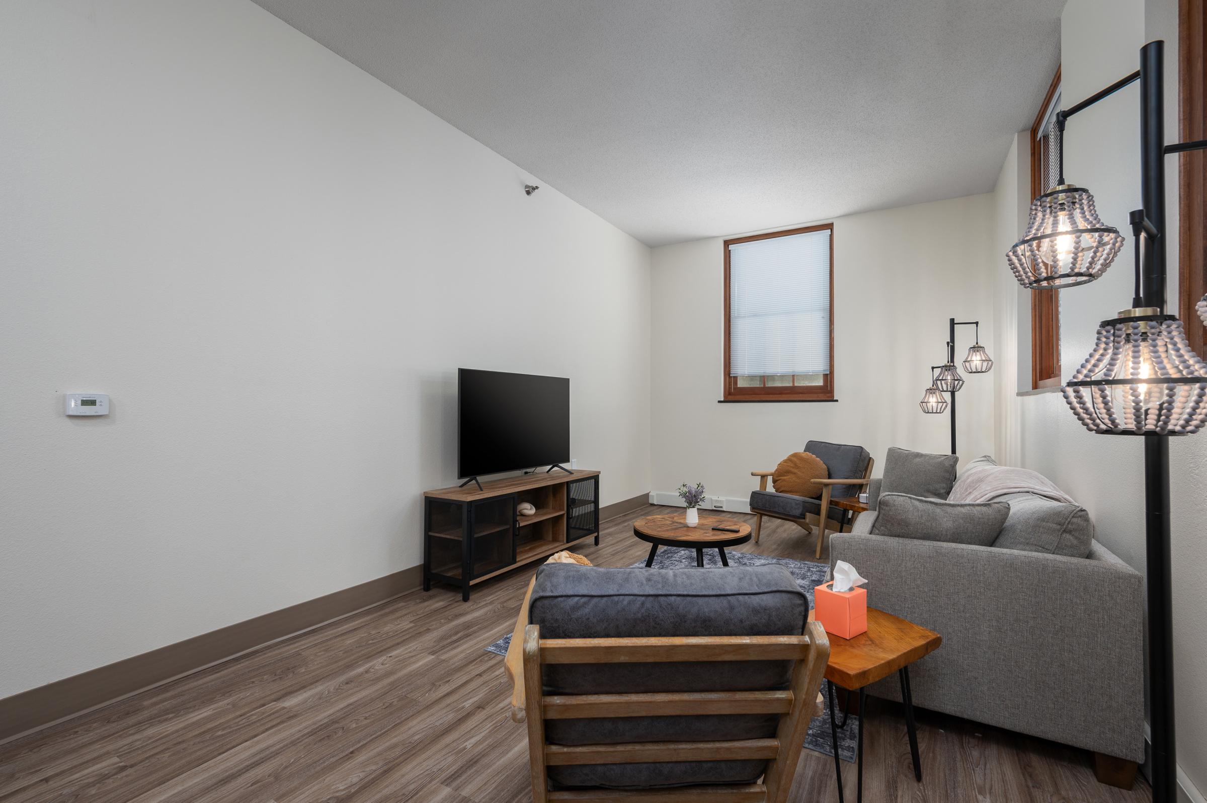 Spacious, modern living room featuring a television, two lounge chairs, a gray sofa, and a round coffee table. Tasteful décor includes lamps and a small tissue box on a side table. Natural light streams in through a window, illuminating the warm wood floor and neutral wall colors.