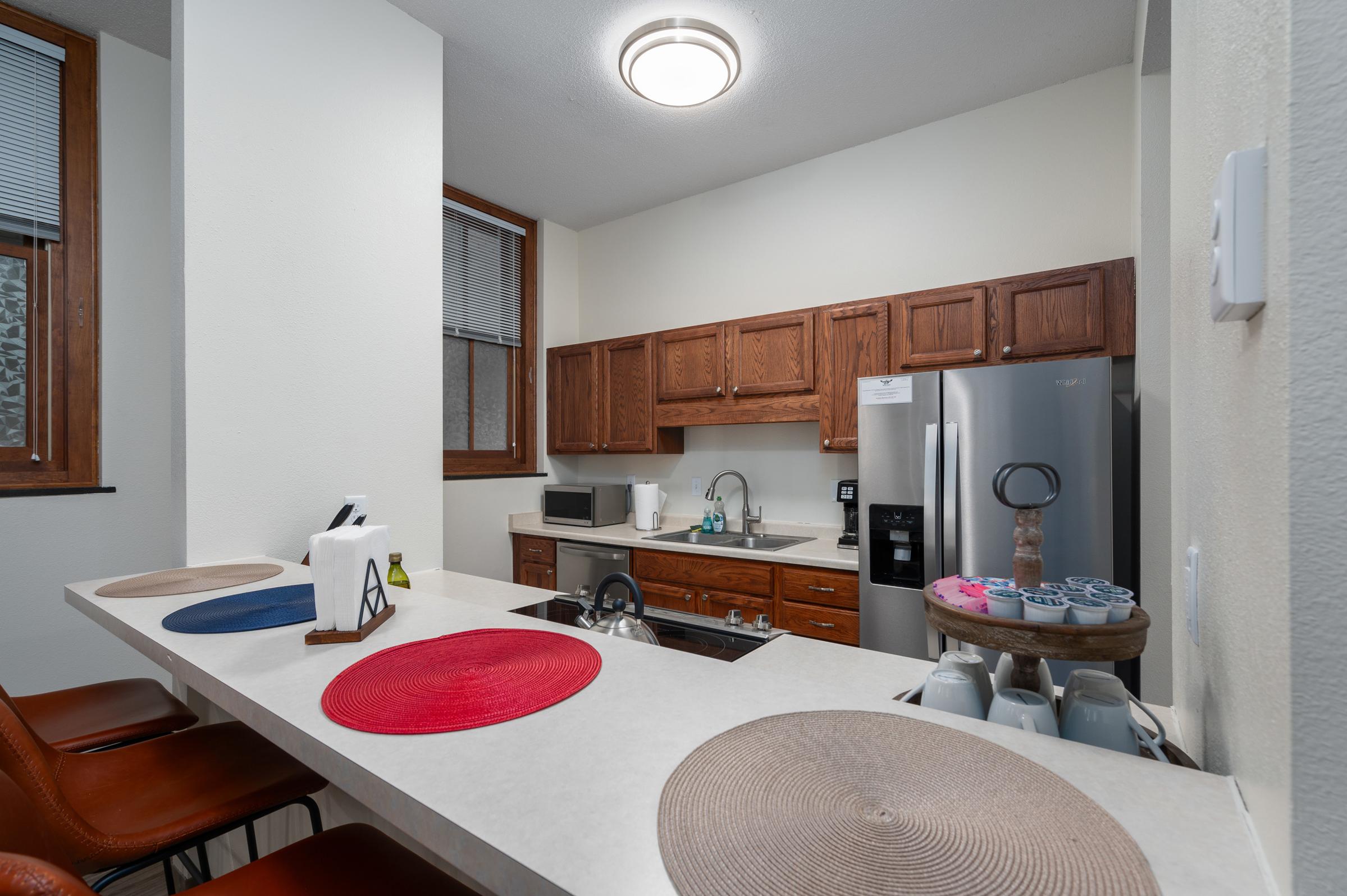 A modern kitchen featuring wooden cabinets, a stainless steel refrigerator, and a countertop with decorative placemats. The space includes a sink, microwave, and an assortment of kitchenware. Natural light enters through a window, illuminating the clean and functional design.