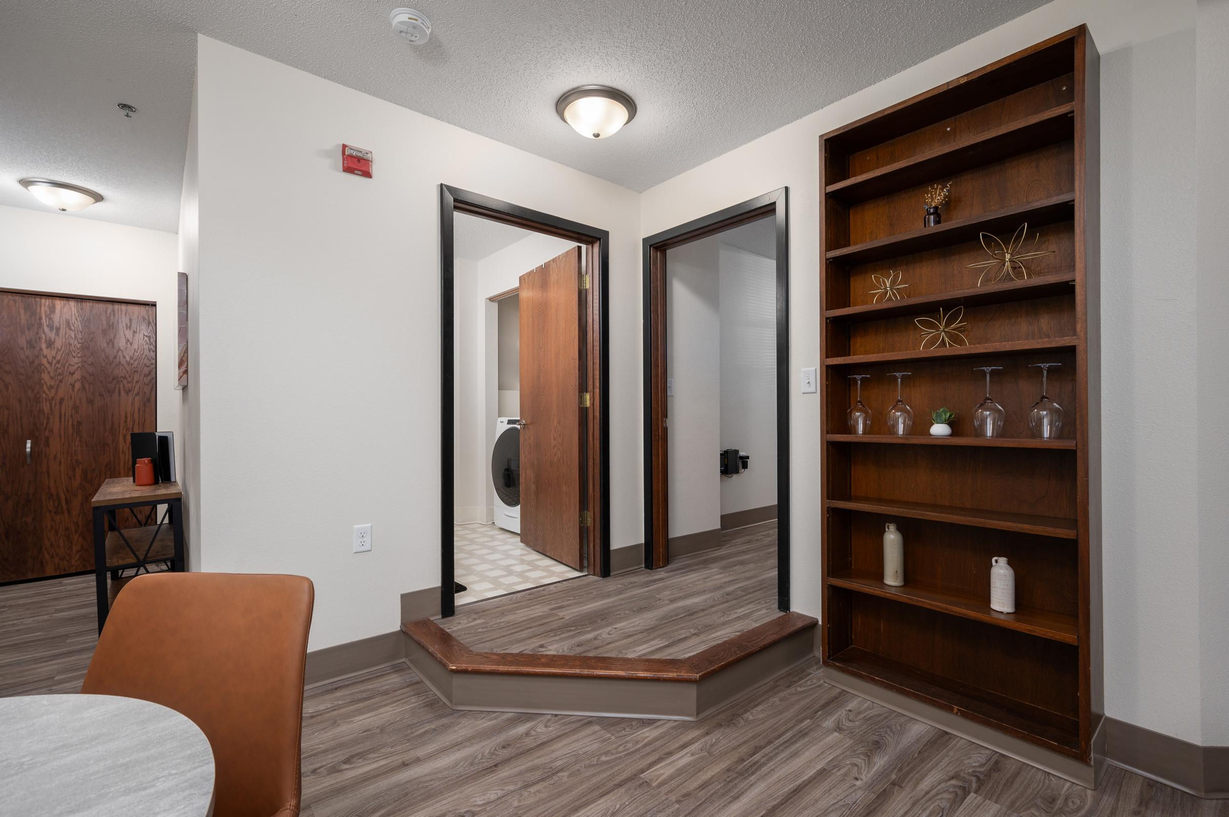 A well-lit interior space featuring a small table with a chair, a bookshelf against the wall, and a staircase leading to another area. Doorways lead to the laundry room and other rooms. The flooring is wood-like laminate, and the walls are painted in light colors.