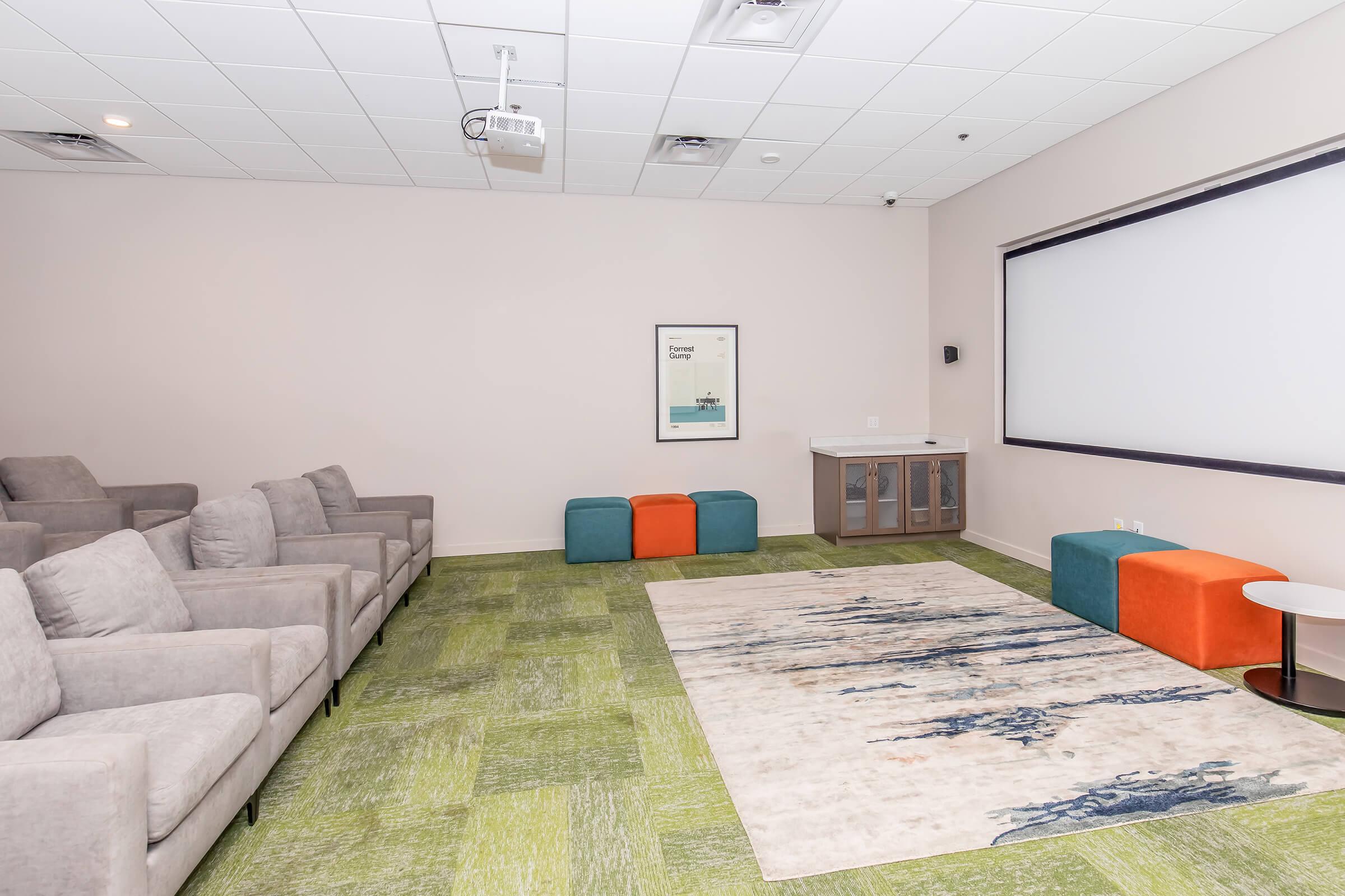 A modern meeting room featuring gray seating, a large projector screen, and a colorful area rug. There are two vibrant benches in orange and teal, along with a side table. The walls are painted a soft beige, and there is a framed artwork mounted above a low cabinet. The carpet is green with a textured pattern.