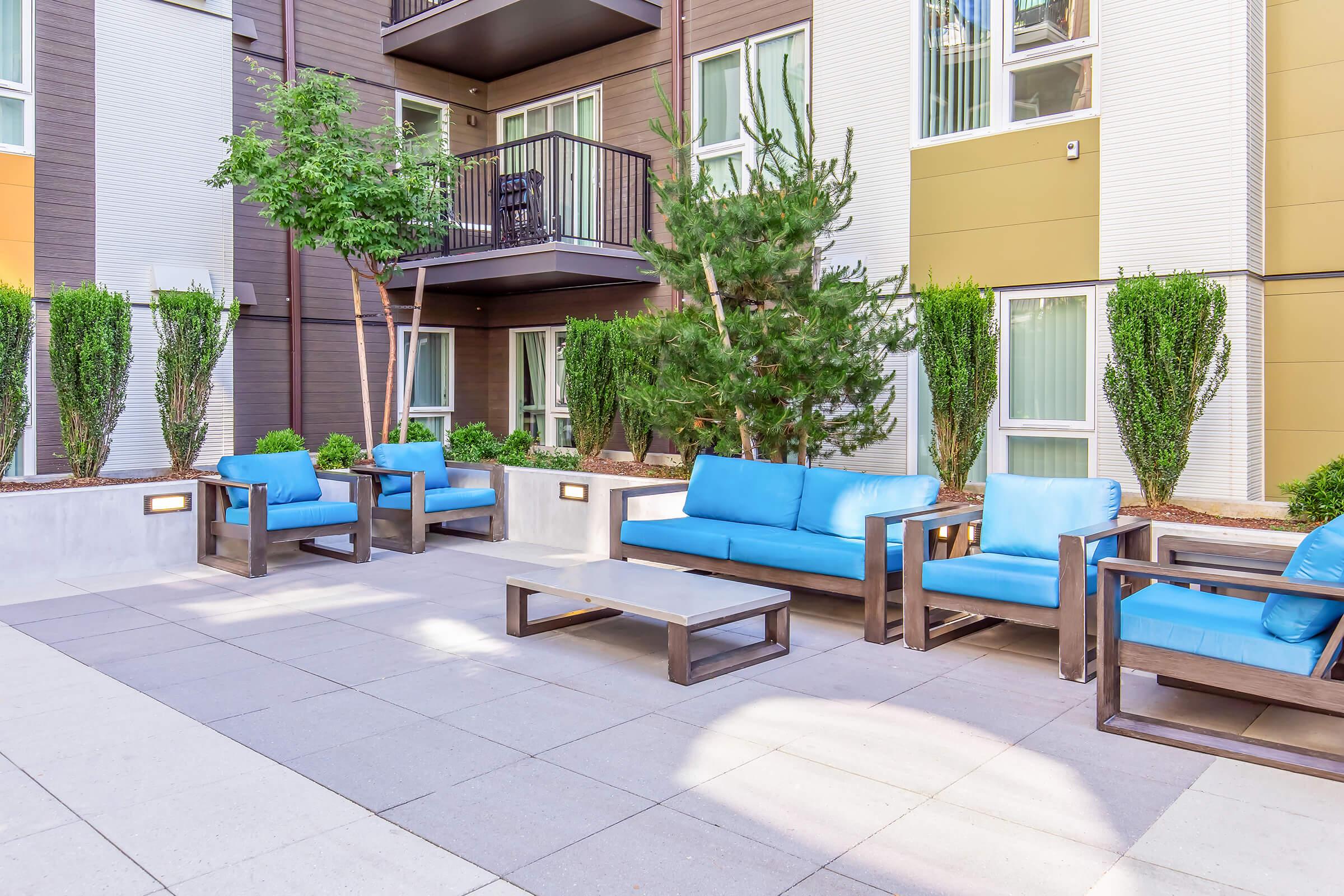 A modern outdoor lounge area with blue couches and a wooden coffee table, surrounded by neatly trimmed plants and trees. The backdrop features multi-story buildings with large windows and balconies, creating a relaxed and inviting atmosphere.