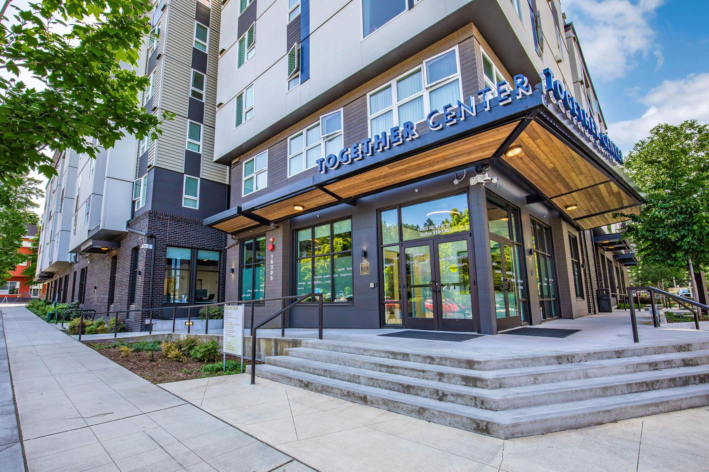 Modern building exterior featuring a welcoming entrance with large glass doors and the sign "Together Center." The surrounding area includes landscaping, walkways, and trees, creating a pleasant urban environment. The architecture showcases a blend of contemporary design elements.
