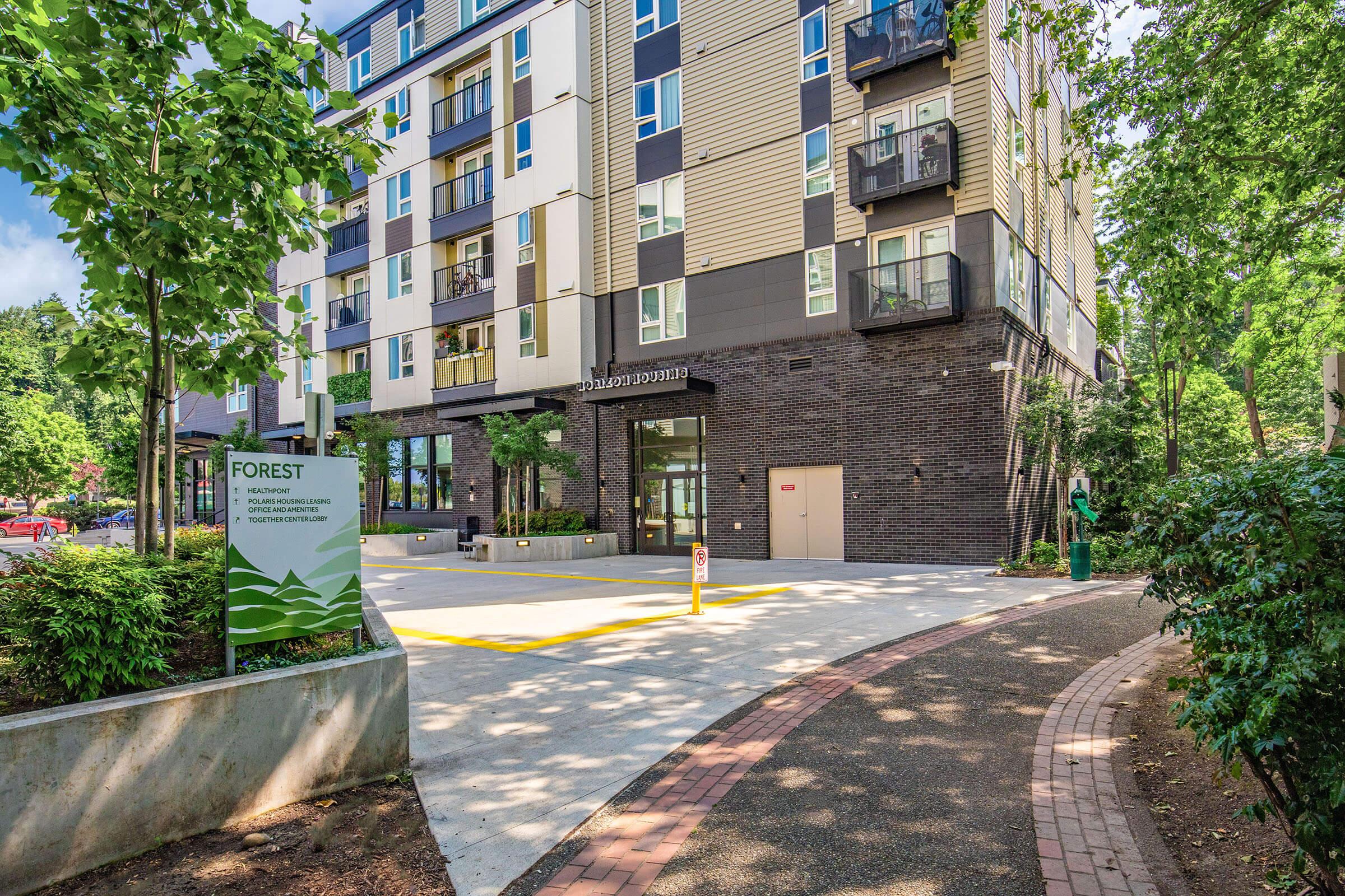 A modern multi-story residential building with a mix of brick and wood facade, surrounded by green landscaping and pathways. The entrance is marked by large windows and a sign for "Forest." The area features trees and shrubs, creating a welcoming outdoor environment.