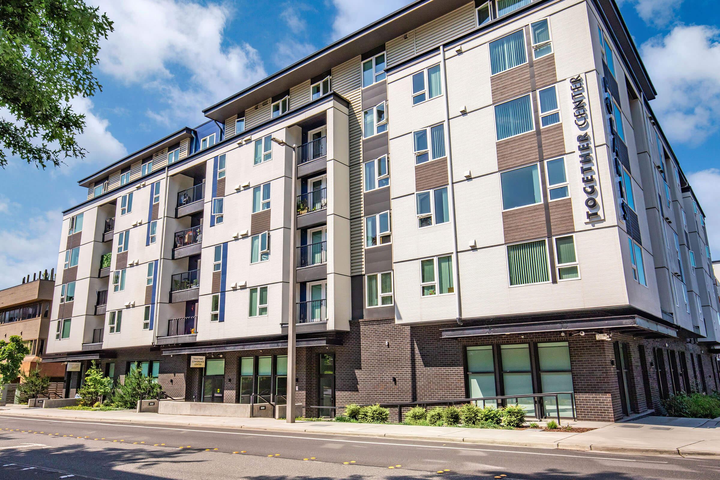 A modern multi-story apartment building with a mix of white and dark exterior panels. Large windows are visible on each floor, and the ground level features storefronts. Lush greenery surrounds the entrance, and the sky is clear with a few clouds.
