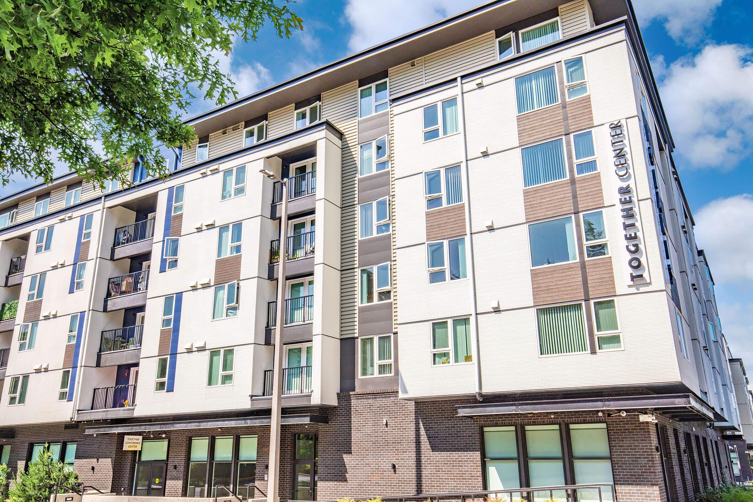 A modern multi-story residential building featuring a mix of white and dark grey exterior. Balconies are visible on several floors, and a sign reading "Together Center" is prominently displayed. The sky is partly cloudy, adding to the daytime ambiance of the scene.