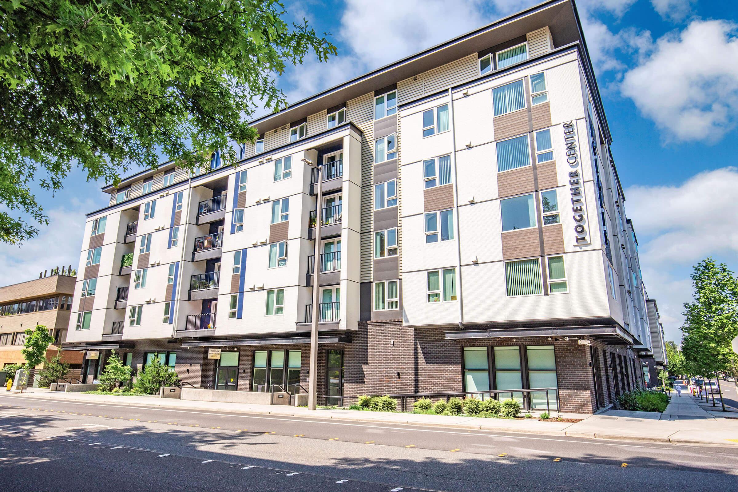 Modern multi-story apartment building with a mix of white and dark brick exteriors. The building features large windows and balconies on the upper floors. Surrounding greenery and trees enhance the urban environment, and the scene is set on a sunny day with blue skies and fluffy clouds.