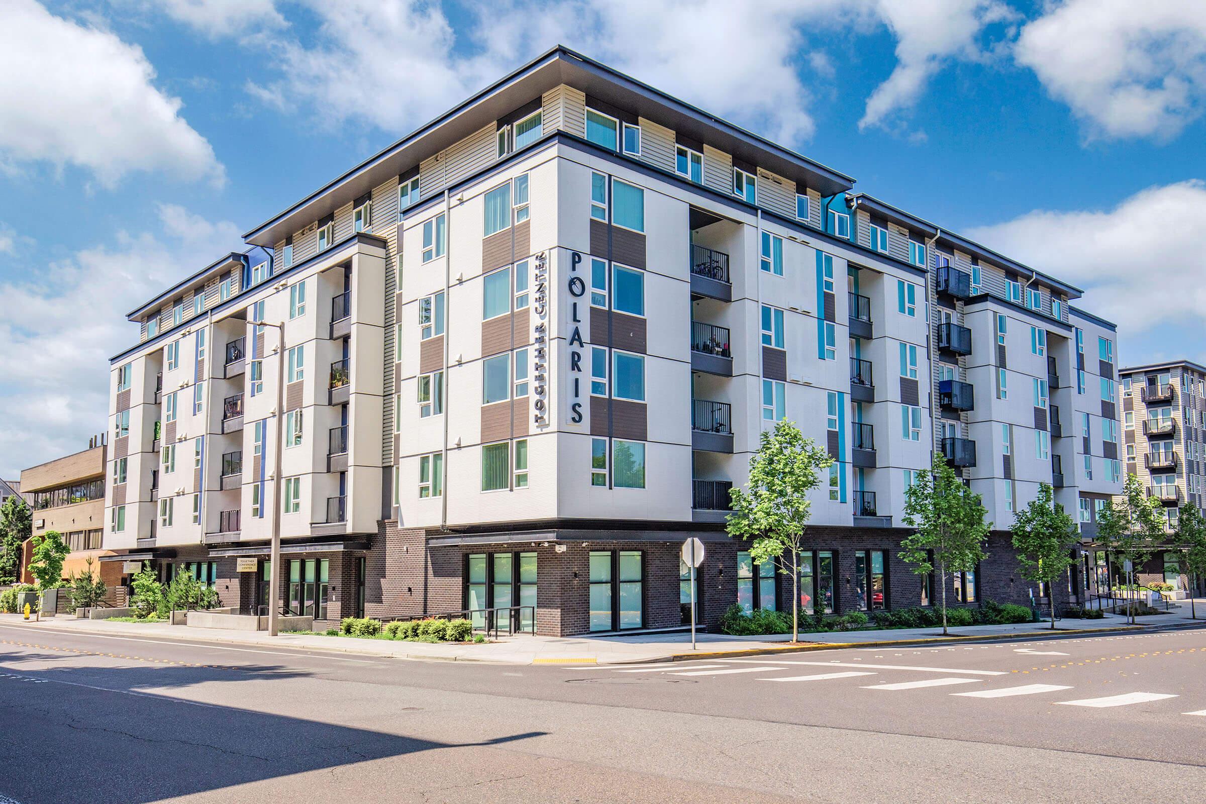 Modern multi-story apartment building with the name "Polaris" prominently displayed. The structure features a combination of white and dark exterior panels, large windows, and balconies. Surrounding the building are landscaped areas with trees and a clear blue sky. Street signs indicate a busy urban environment.
