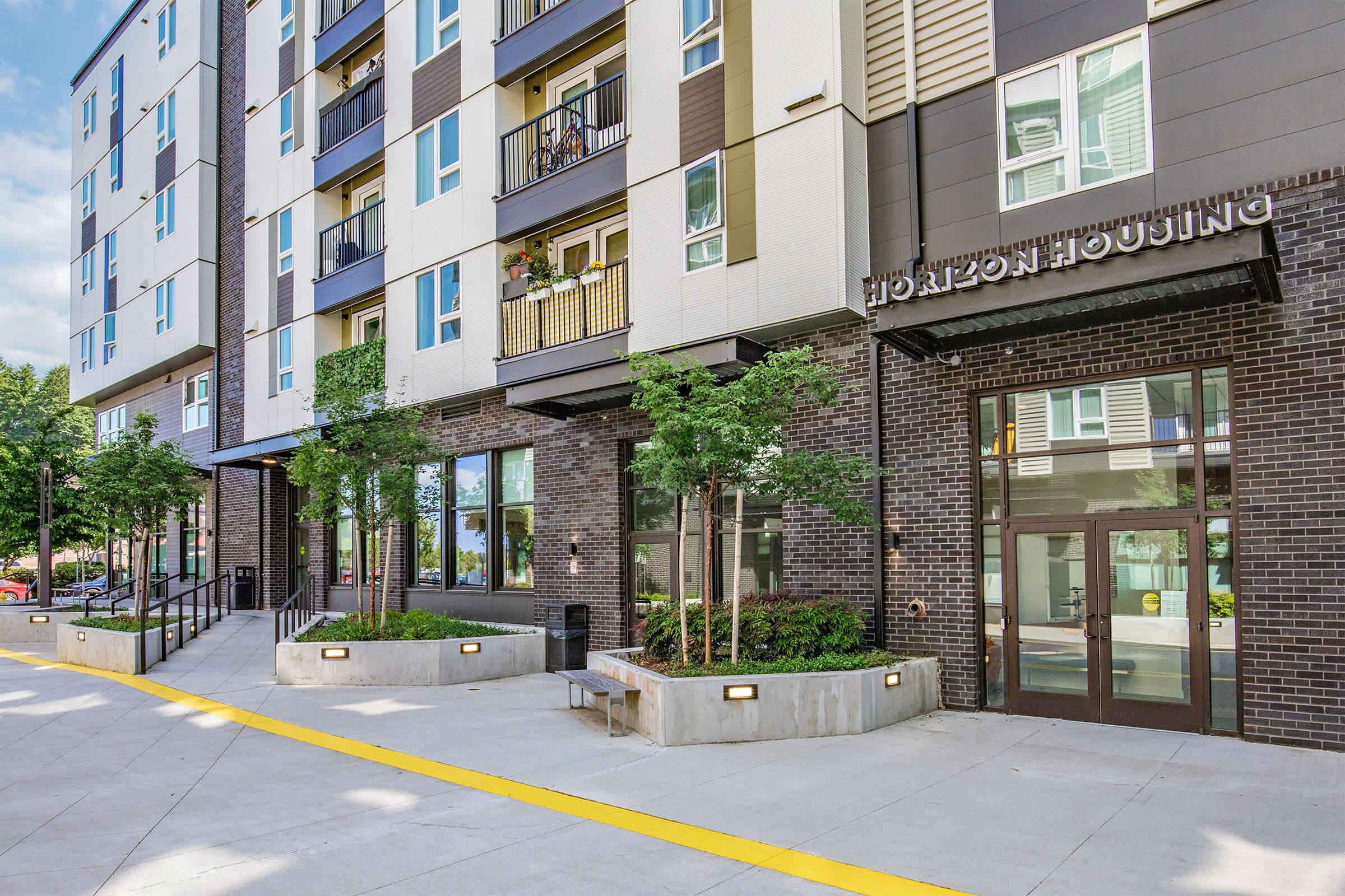 Modern apartment building with a brick facade and glass doors. The entrance features a sign that reads "Horizon Housing." Surrounding the entrance are small trees and landscaped areas, with a wide yellow pathway leading to the door. The sky is clear, and the setting appears vibrant and welcoming.