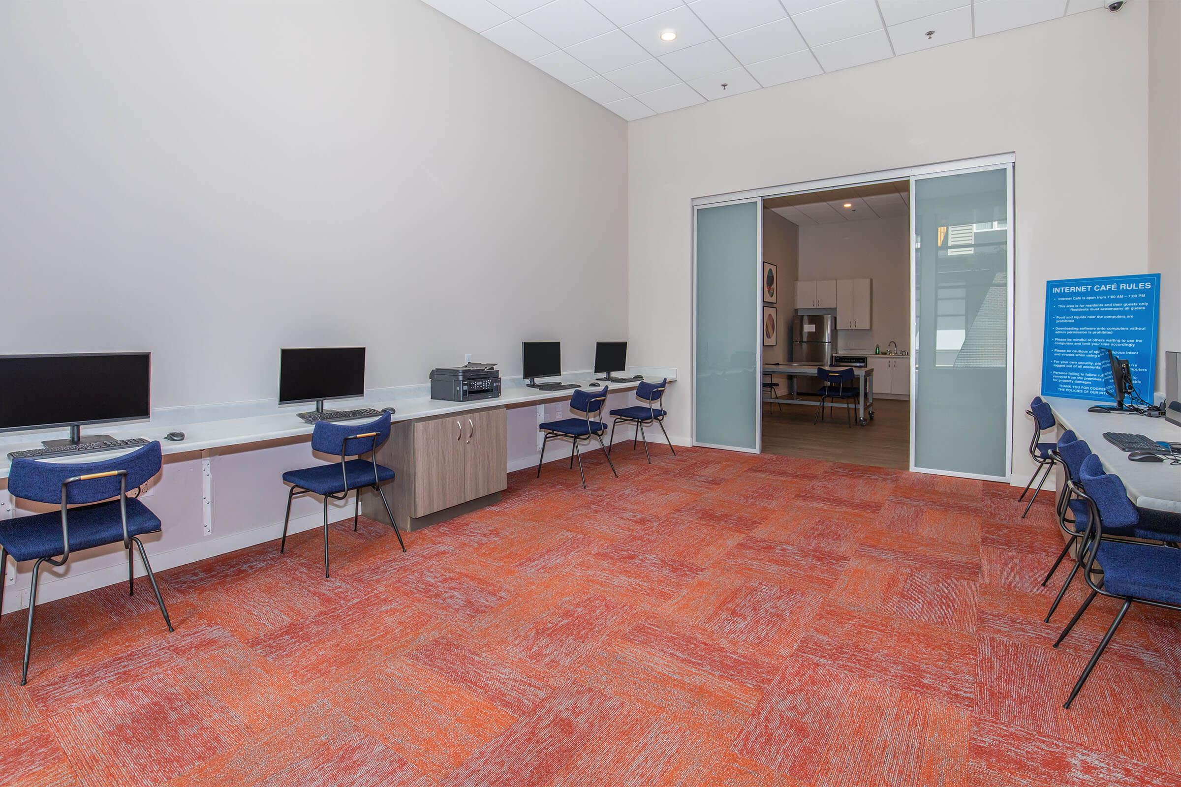 A modern workspace featuring several computers on desks along the walls, with a spacious area and orange carpet flooring. The room has large glass doors leading to another area, and a blue informational sign on the right. Comfortable chairs are arranged at the computer stations.