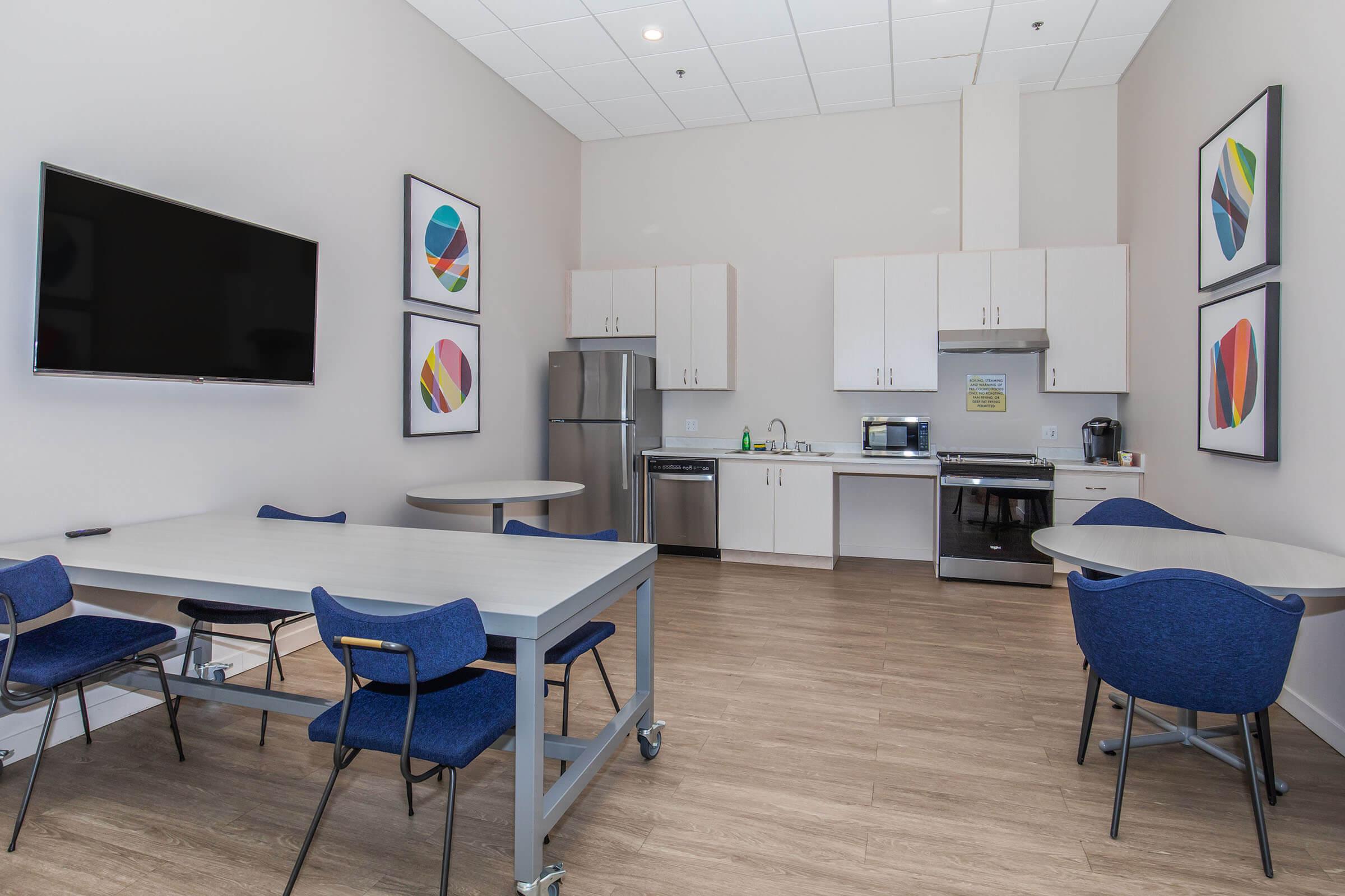 A modern kitchen area featuring a large flat-screen TV, a refrigerator, a microwave, and a stove. There are several tables with blue chairs and colorful abstract art on the walls, creating a bright and inviting atmosphere for meals or meetings.