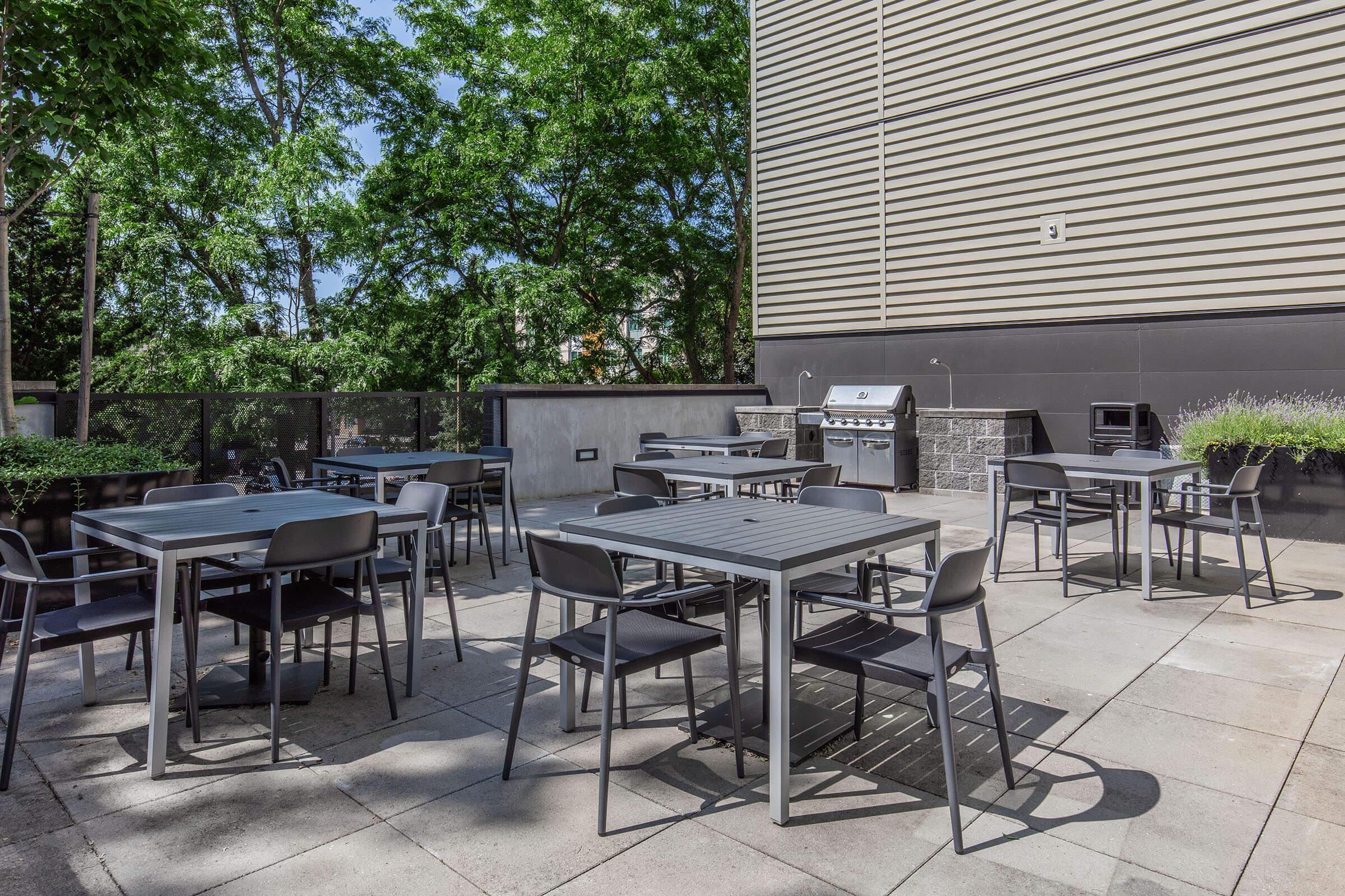 Outdoor seating area featuring several gray metal tables and chairs on a tiled patio. Lush green trees provide shade in the background, and a barbecue grill is positioned against the wall. The setting is bright and inviting, ideal for gatherings or meals outside.