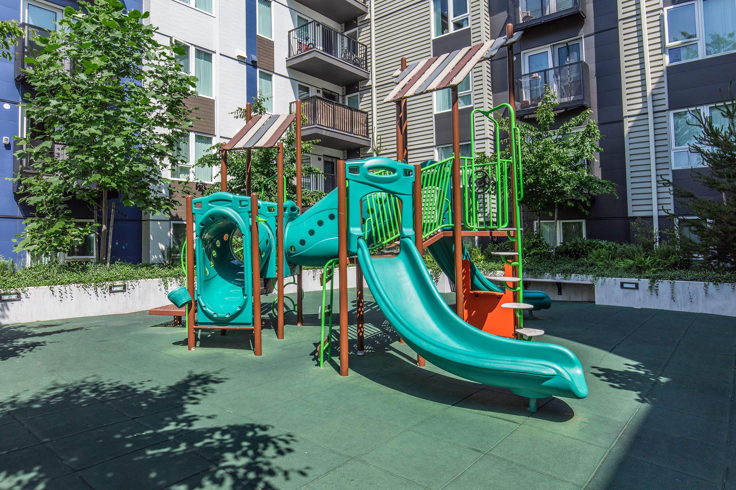 Colorful playground equipment featuring a slide and climbing structures, surrounded by trees and modern apartment buildings. The play area is designed with safety flooring and includes shaded areas, creating a welcoming environment for children.