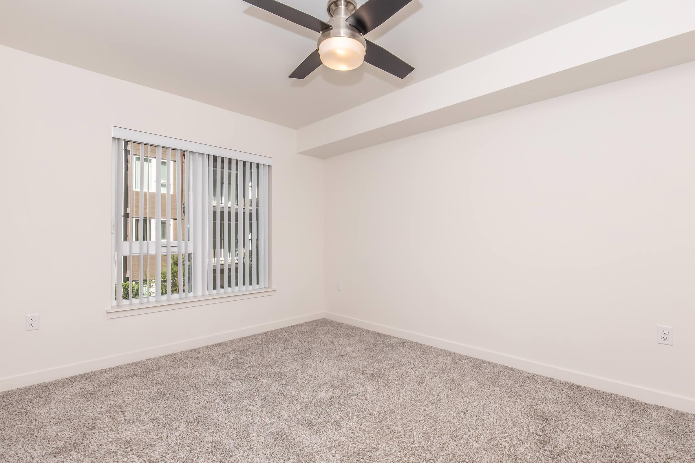 A spacious, empty room with light beige walls and a plush carpet. It features a ceiling fan with a light fixture and a window with vertical blinds allowing natural light to enter. The room has a clean, minimalistic design, making it suitable for various purposes such as a bedroom or office.