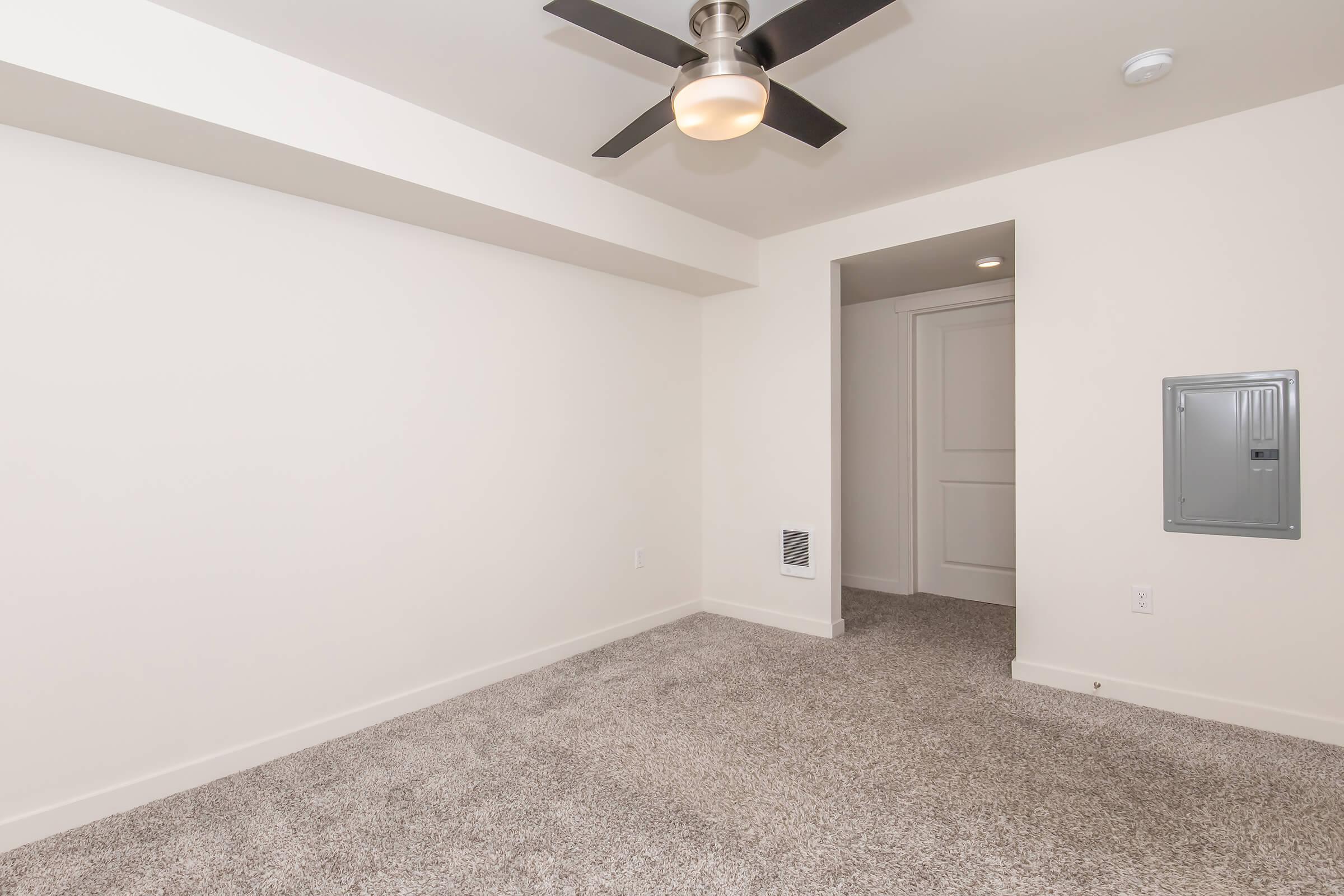 A spacious, empty room with light beige walls and grey carpet flooring. It features a ceiling fan, a doorway leading to another area, and an electrical panel on the wall. The overall aesthetic is modern and minimalistic, ideal for customization.