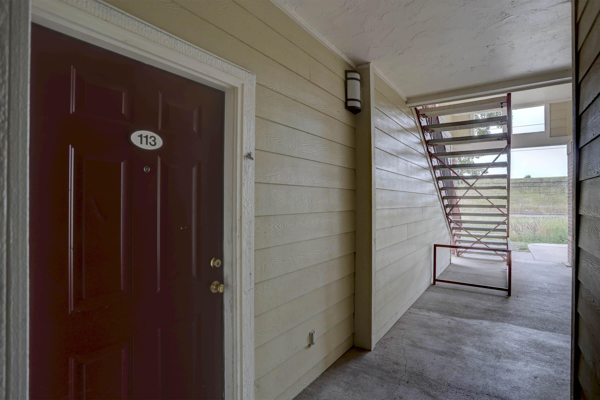 View of an apartment entrance with a brown door labeled "113." The hallway features light-colored siding, a metal staircase leading to another level, and an outdoor view with greenery in the background. The floor is concrete, and natural light illuminates the area.