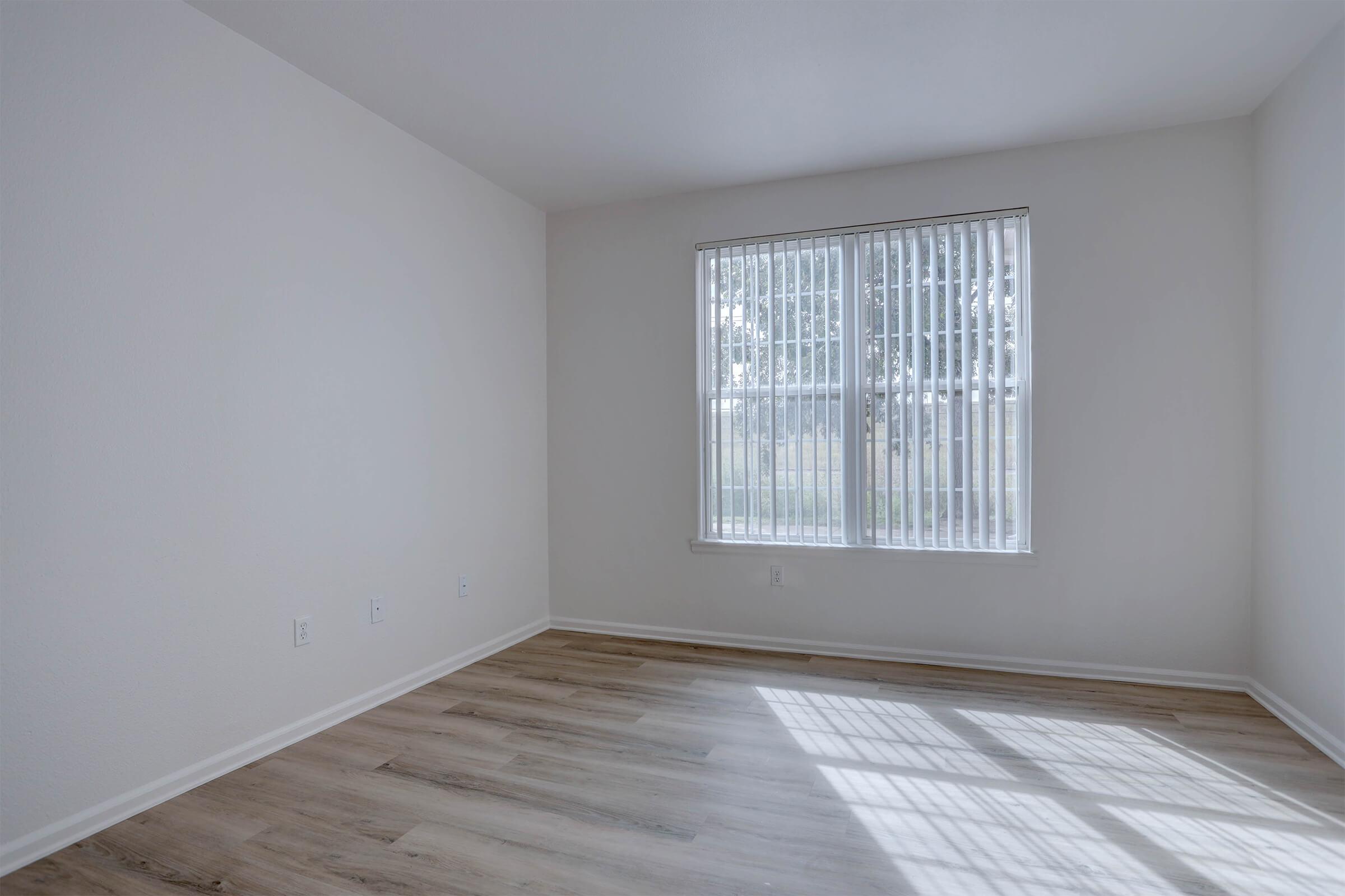 A light-filled, empty room with a large window featuring vertical blinds. The floor is wooden, and the walls are painted a soft white. Sunlight casts shadows on the floor, highlighting the spaciousness of the room.