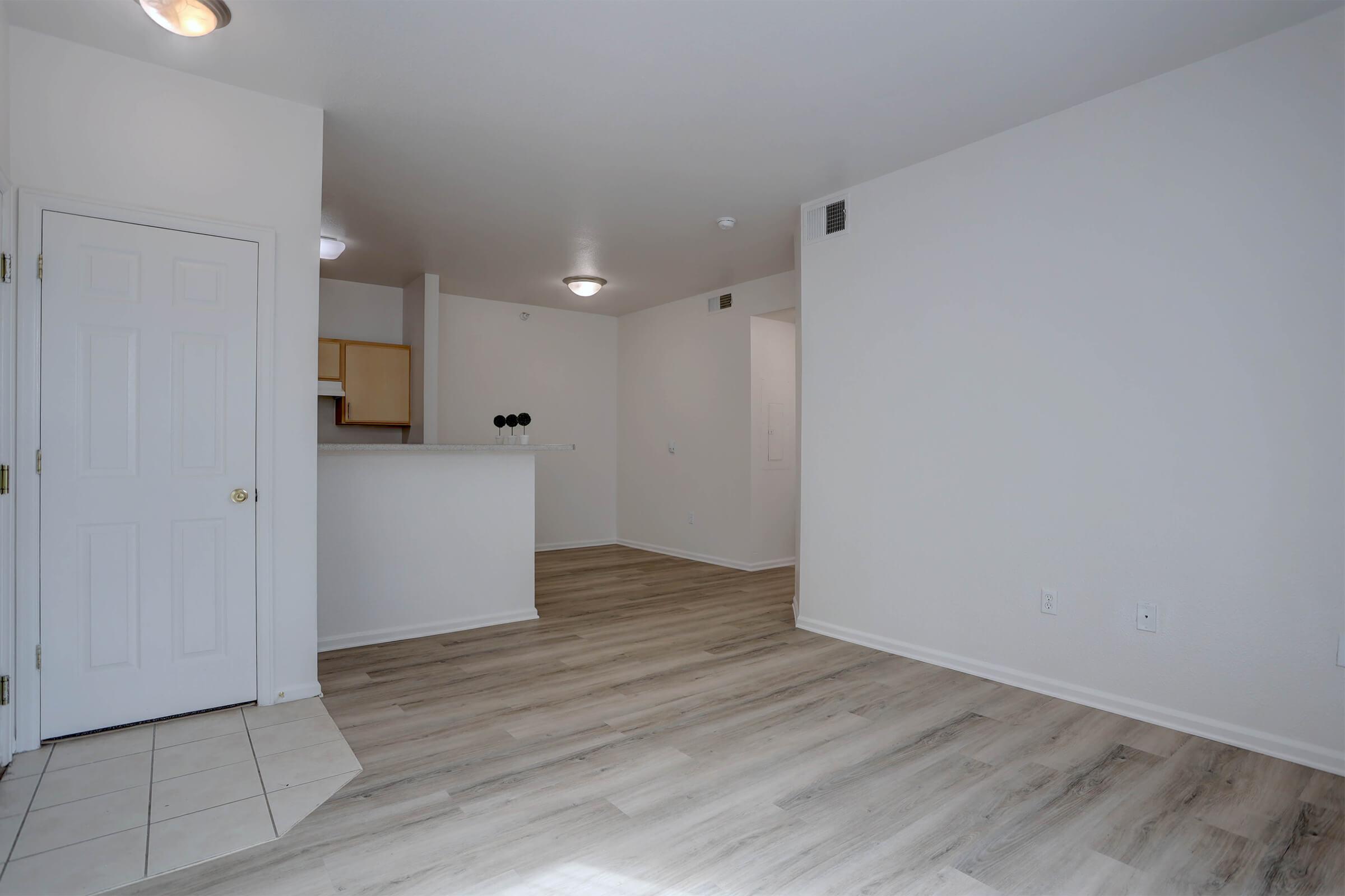 Interior view of a spacious, empty living area with wood-like flooring. A kitchen area is visible in the background, featuring light cabinetry. The walls are painted in a soft, neutral color, and there are light fixtures on the ceiling. The scene is well-lit, creating an inviting atmosphere.