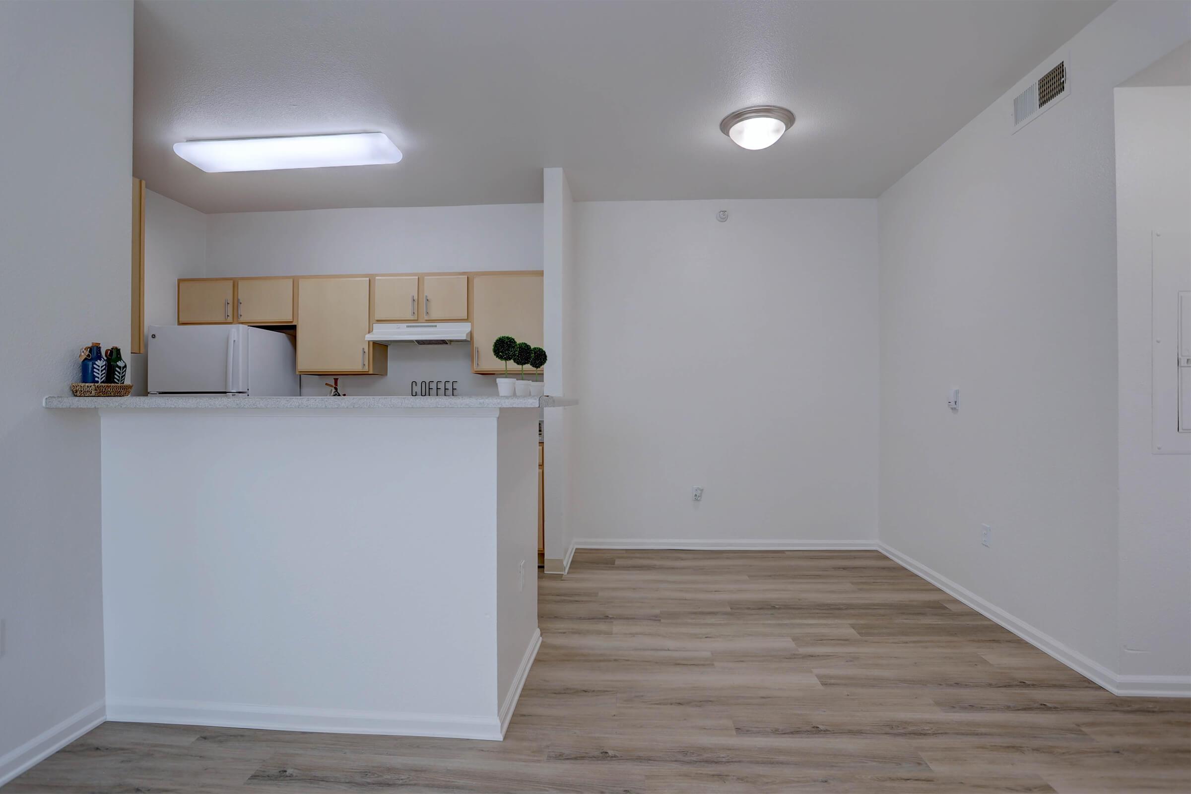 A modern kitchen and living area with light wood flooring. The kitchen features beige cabinets, a white refrigerator, and a countertop with decorative items. The space is bright and airy, with neutral-colored walls and a ceiling light fixture.