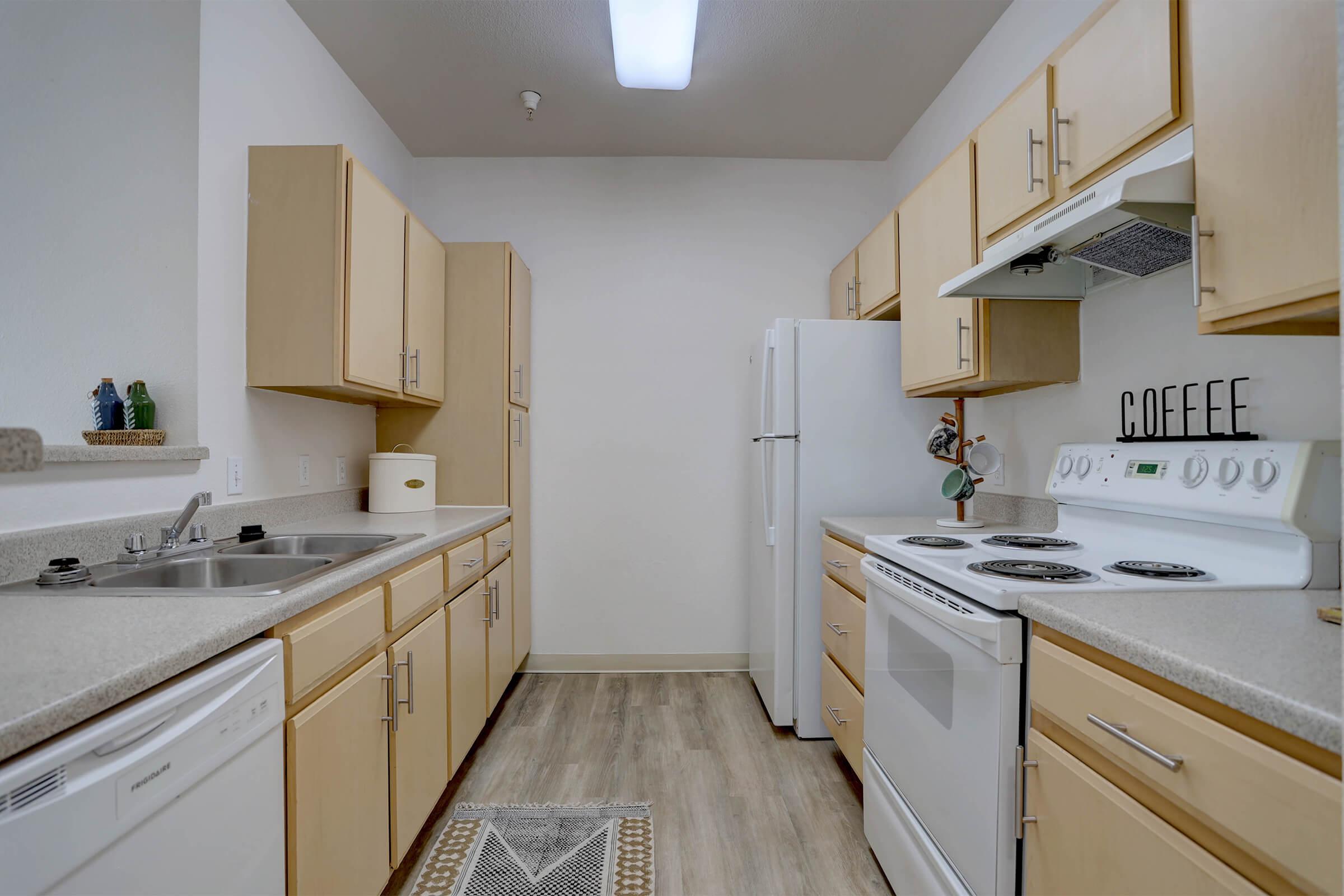 A modern kitchen featuring light wood cabinets, a white stove, refrigerator, and dishwasher. The countertops are light-colored, and there's a small rug on the floor. A coffee-themed decoration is on the wall, and the overall ambiance is bright and functional.