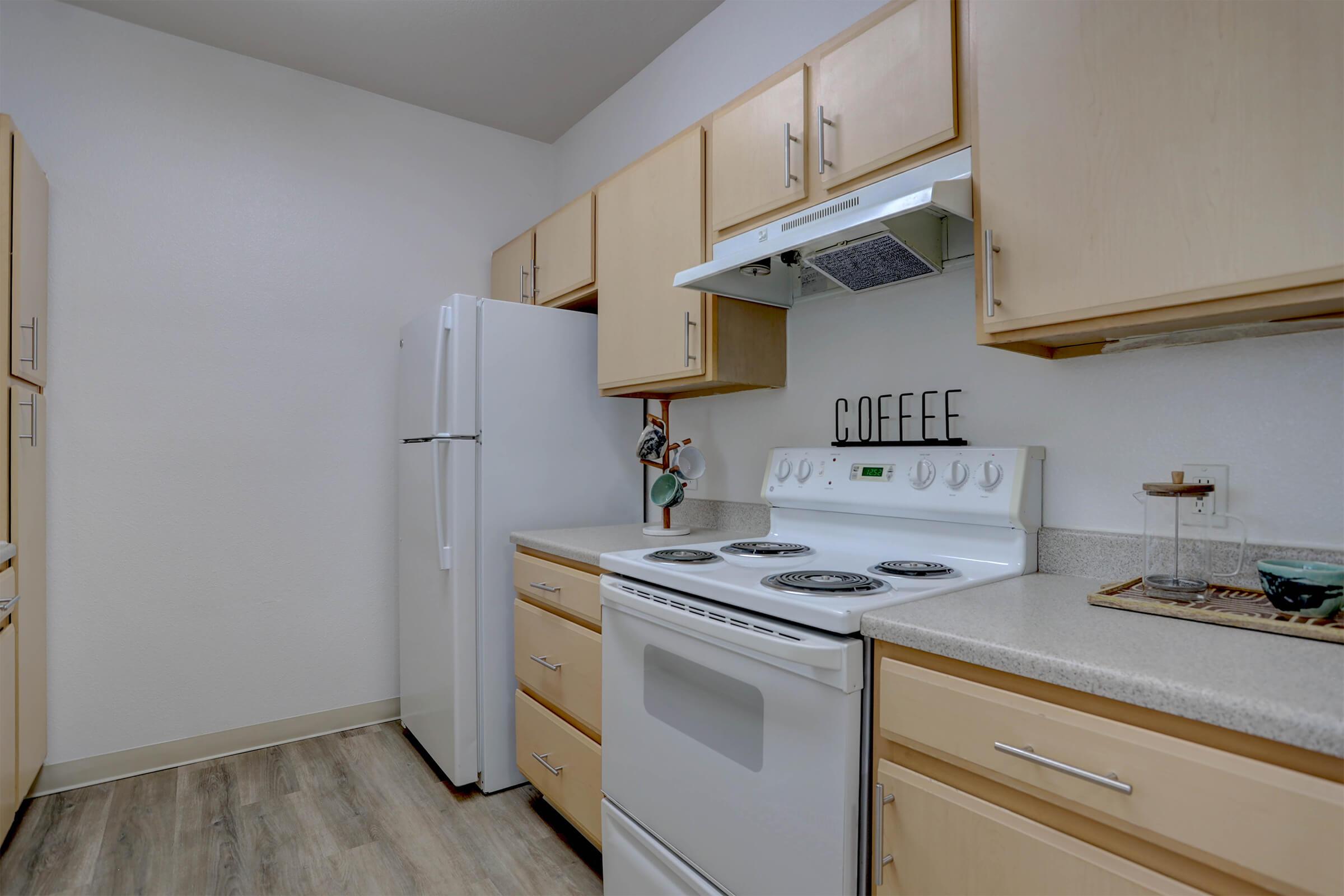 A modern kitchen with light wood cabinets, a white stove, and a refrigerator. The counter features a coffee station with a container labeled "COFFEE" and a plant. The room has light-colored walls and laminate flooring, creating a bright and inviting atmosphere.