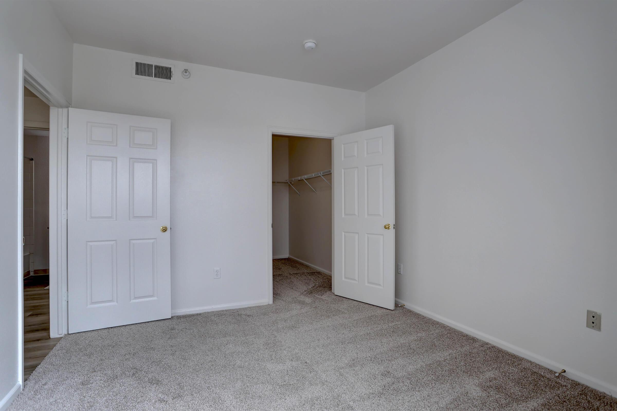 Empty room with light-colored walls and plush carpeting. Two open doors lead to separate spaces, one with a closet featuring a hanging rod. The natural light illuminates the space, creating a bright and airy atmosphere. The room appears freshly painted and well-maintained.