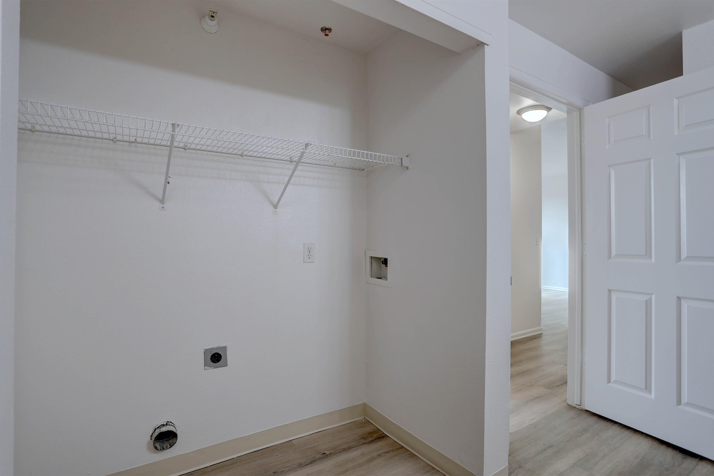 Empty laundry room space with a wall-mounted wire shelf, a wash machine outlet on the floor, and a small access panel. A white door is partially open, leading to another room with light-colored flooring. The walls are painted a light shade, creating a clean and spacious appearance.