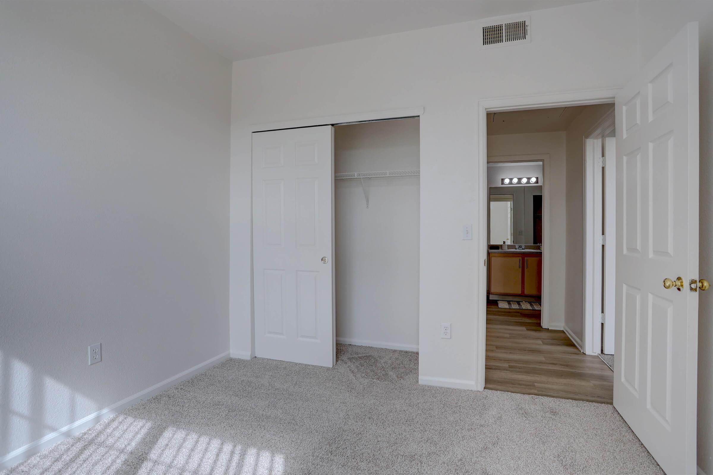A bright, empty bedroom featuring light-colored walls and plush carpet. There is a sliding closet door on the left side, which is open to reveal a closet space. To the right, there is an entrance to a bathroom visible, with a wooden floor and a mirror. Soft natural light filters through the window, creating a warm atmosphere.