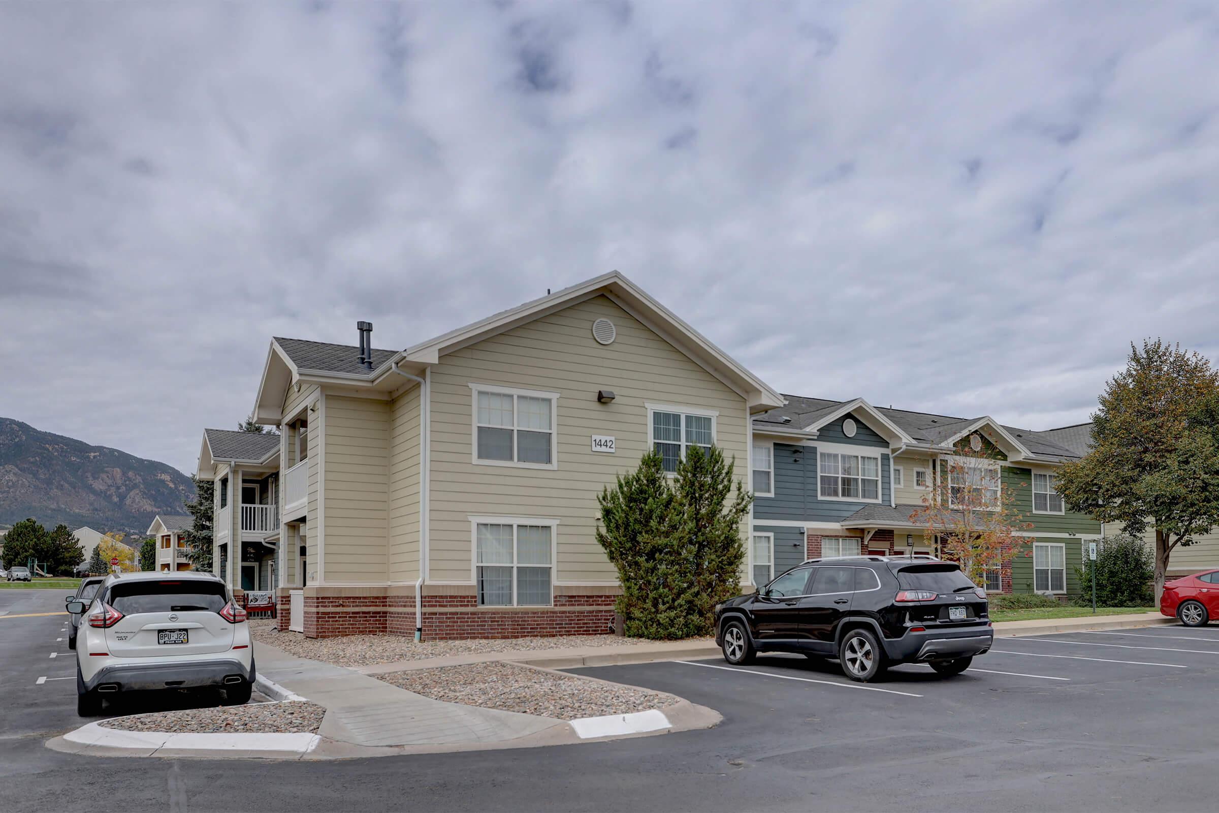 Multistory residential apartment building with a light-colored exterior, surrounded by parking spaces. Several vehicles are parked nearby, including a white SUV and a black car. The sky is overcast, and there are mountains in the background along with trees near the building.