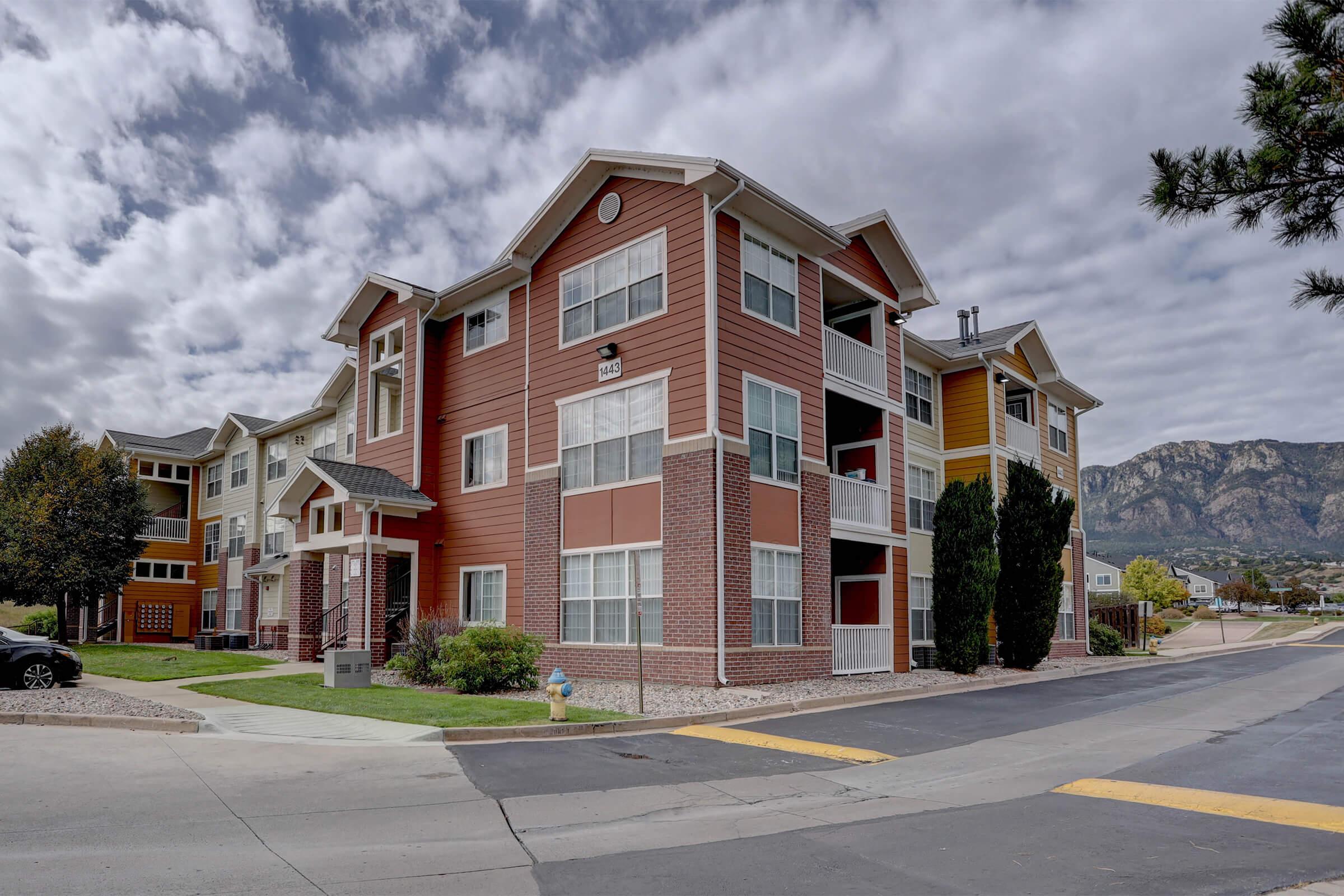 Modern multi-story apartment building featuring a combination of brick and colorful siding. The structure includes large windows, a landscaped area with trees and shrubs, and is situated near a road with mountains in the background under a cloudy sky.