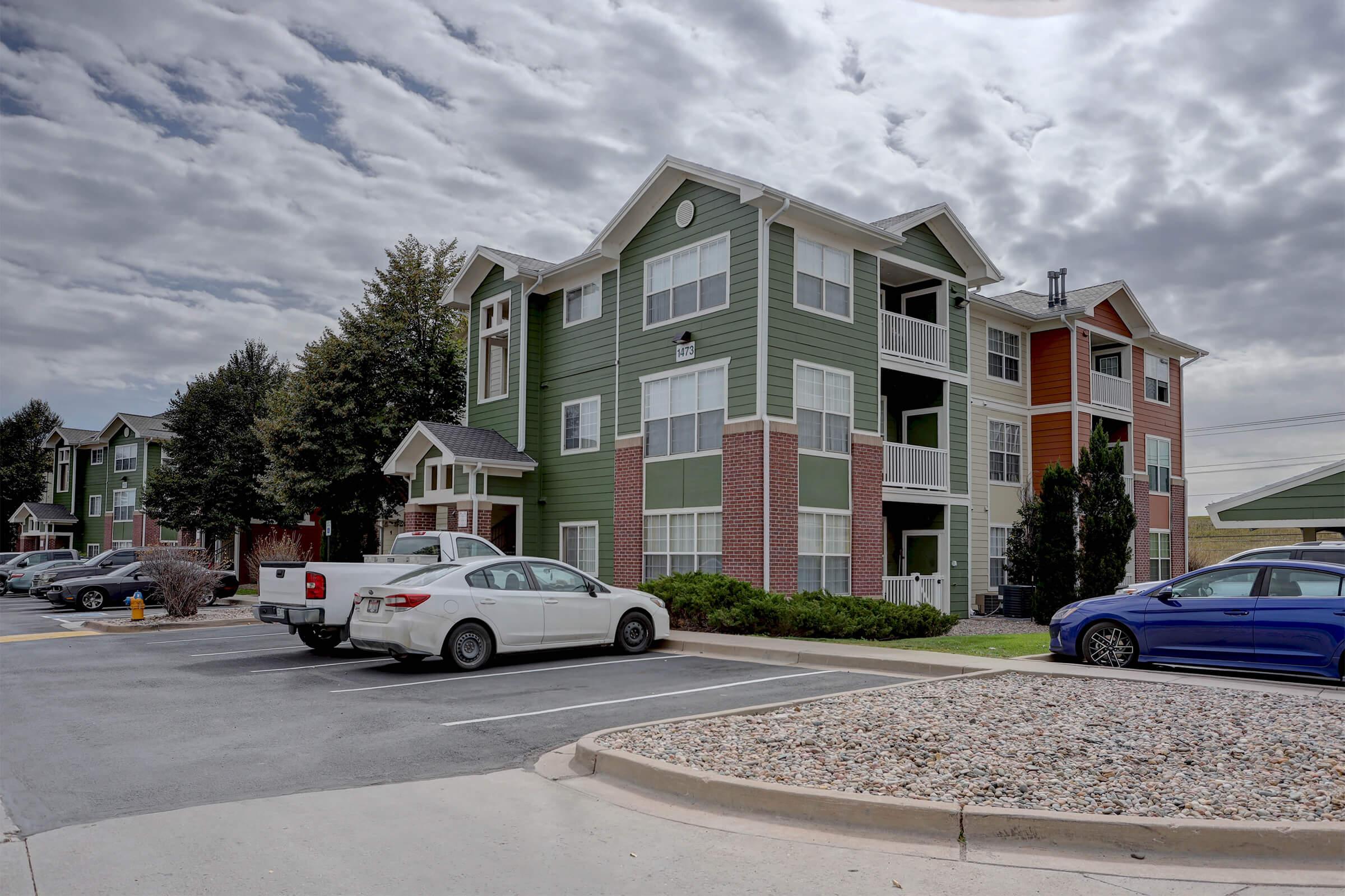 A three-story residential apartment building with a mix of green and orange siding. The building has multiple windows and balconies. In the foreground, there are several parked cars on a paved lot, along with a landscaped area featuring grass and small shrubs. The sky is partly cloudy.