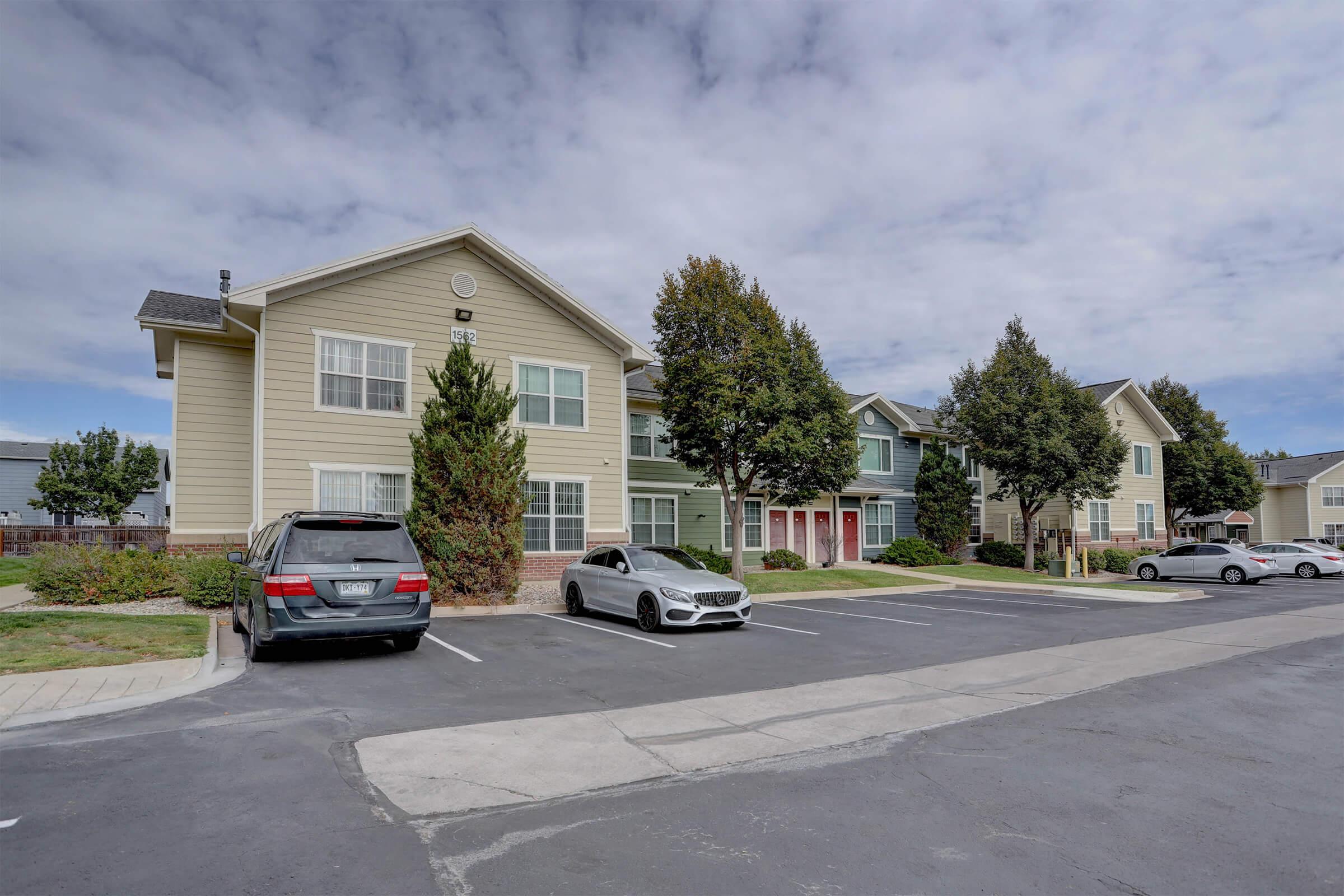 A view of a residential area showcasing two multi-unit buildings with light-colored siding. Several parked cars are visible in front of the buildings, with trees providing greenery. The sky is partly cloudy, creating a pleasant atmosphere in the neighborhood.
