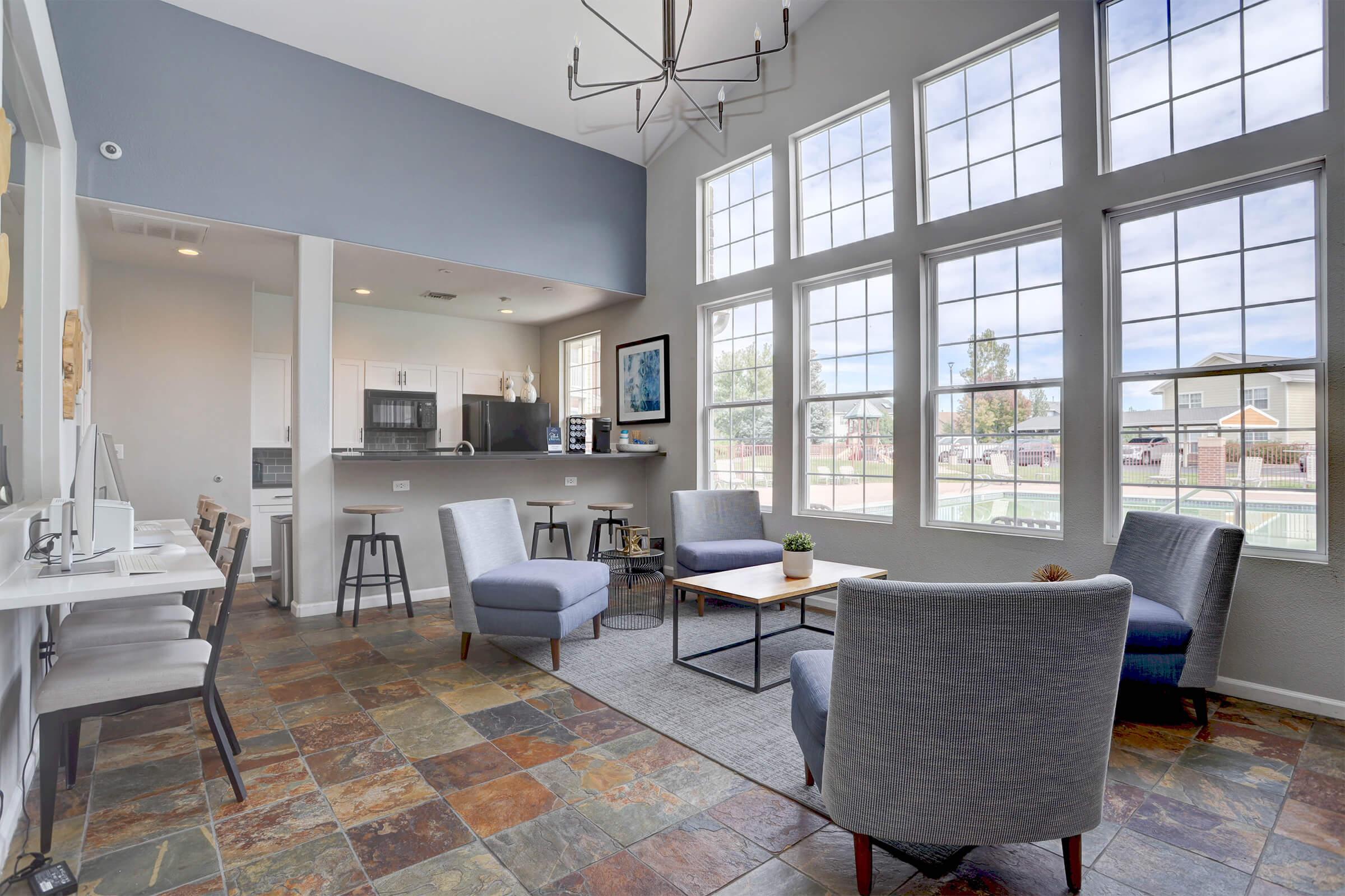 A modern living space featuring large windows that let in natural light. It includes two gray armchairs, a coffee table, and a textured rug on a slate-tiled floor. In the background, there's a kitchen area with bar stools, and colorful artwork adorns the walls. The overall atmosphere is inviting and stylish.