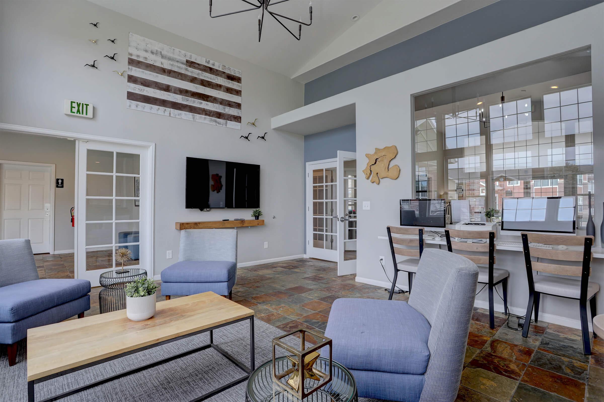 A modern common area featuring grey couches and a wooden coffee table. A wall with a decorative flag and birds is in the background. There's a television mounted on the wall and a work station with two computers in the corner. Natural light floods the space through large windows, creating a welcoming atmosphere.