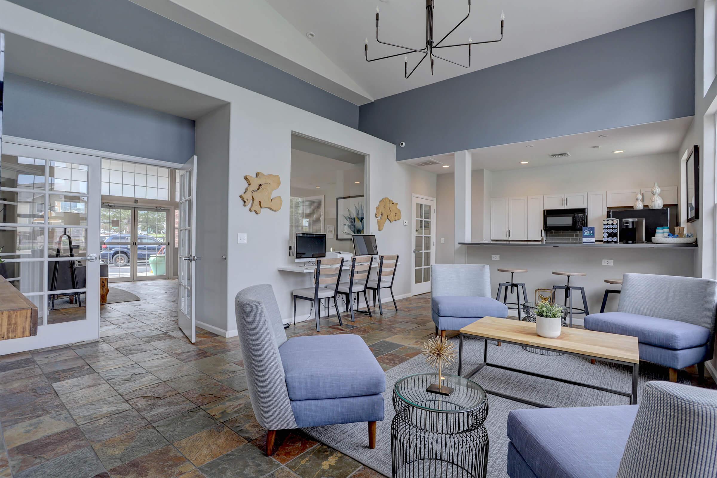 A modern interior space featuring a spacious living area with gray upholstered chairs, a wooden coffee table, and decorative plants. Large windows let in natural light, while a kitchen area with white cabinetry and black appliances is visible in the background. The floor has a stylish slate design.
