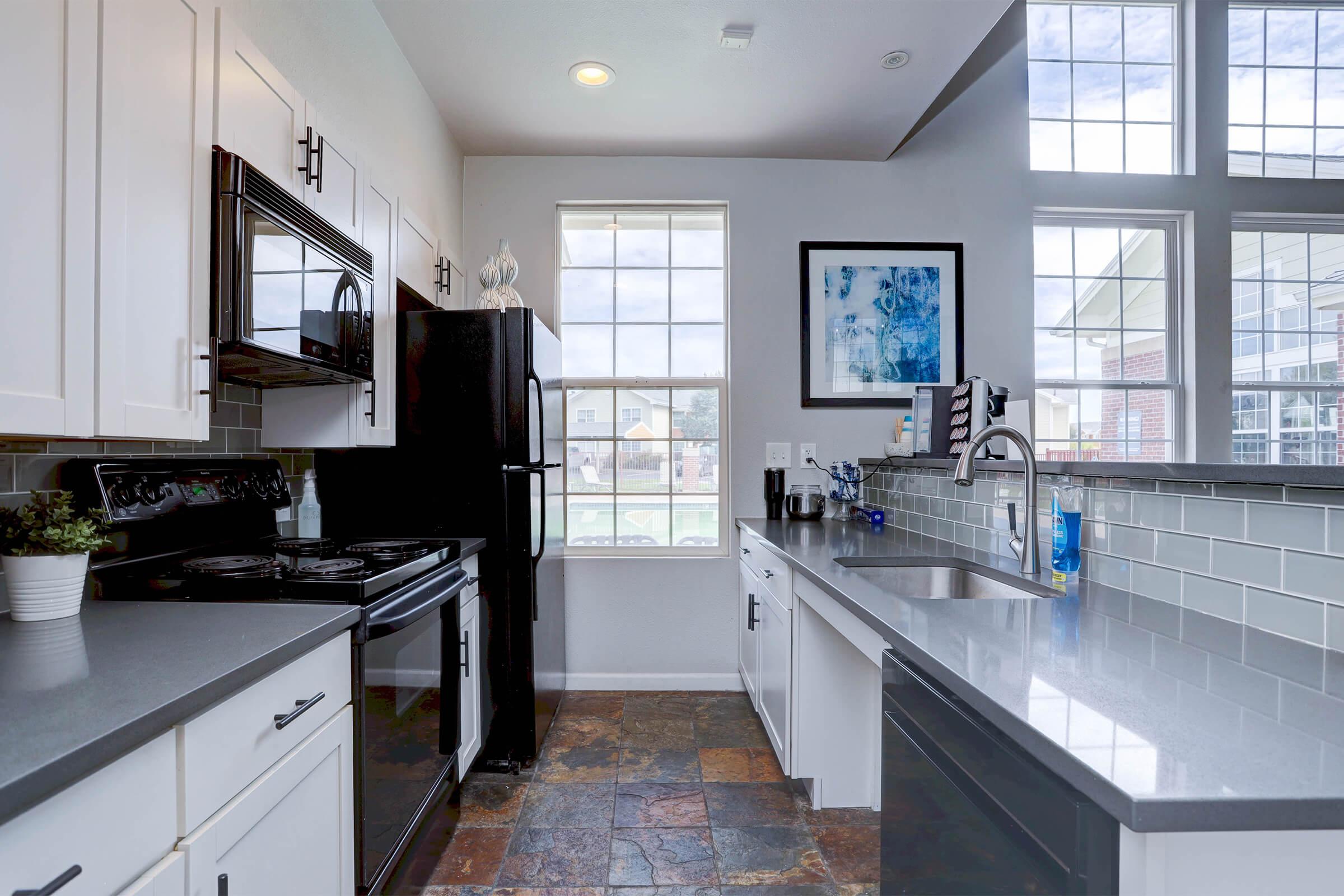 A modern kitchen featuring black appliances, including a microwave and refrigerator, white cabinets, and a gray countertop. Natural light streams in through large windows, creating a bright atmosphere. The floor is made of slate tiles, and there's a small plant on the counter, adding a touch of greenery.