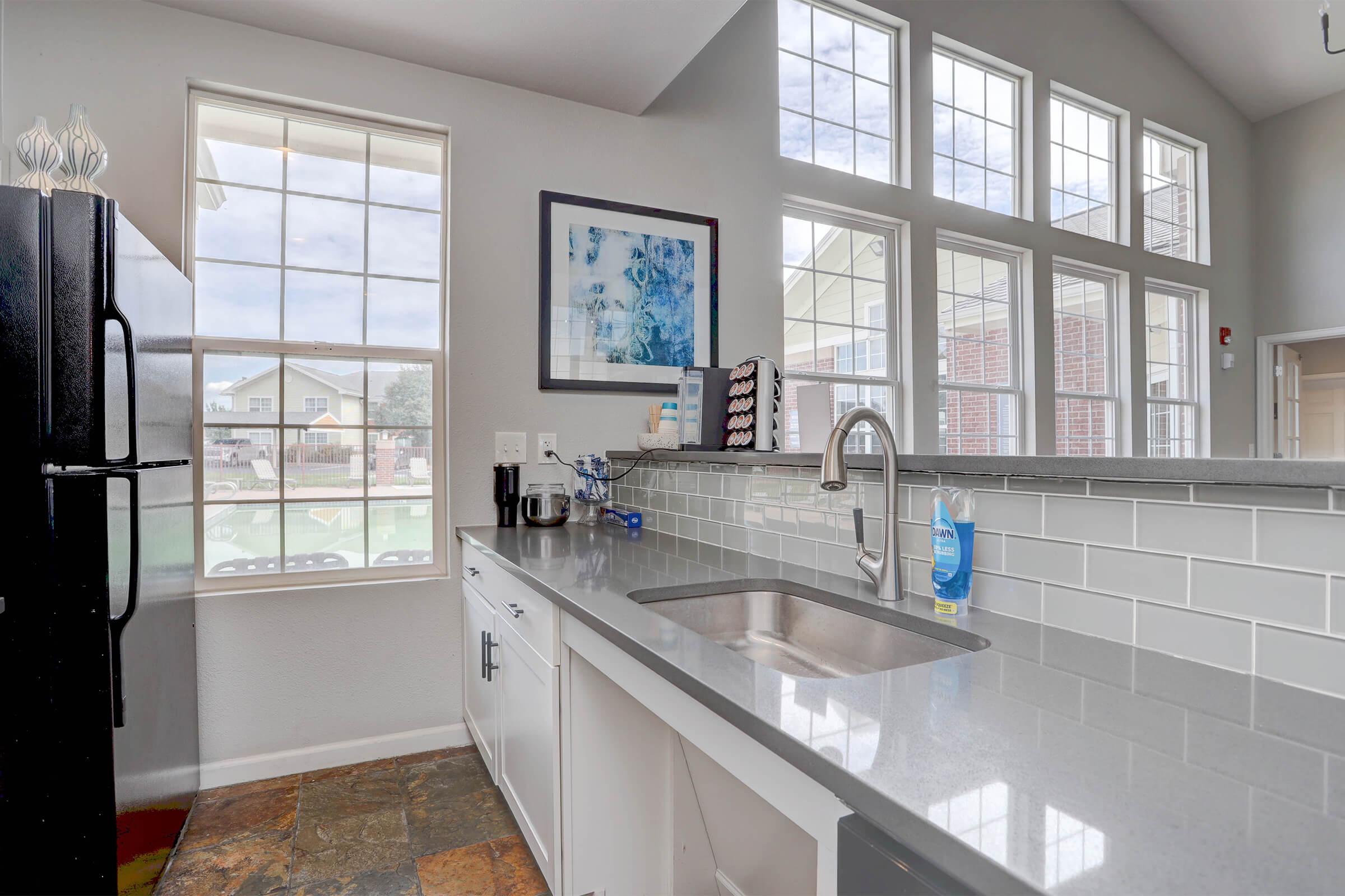 Modern kitchen featuring a sleek countertop, stainless steel sink, and a black refrigerator. Large windows allow natural light, complemented by stylish wall art. The backsplash is decorated with white tiles, enhancing the contemporary design. Overall, the space is bright and inviting.