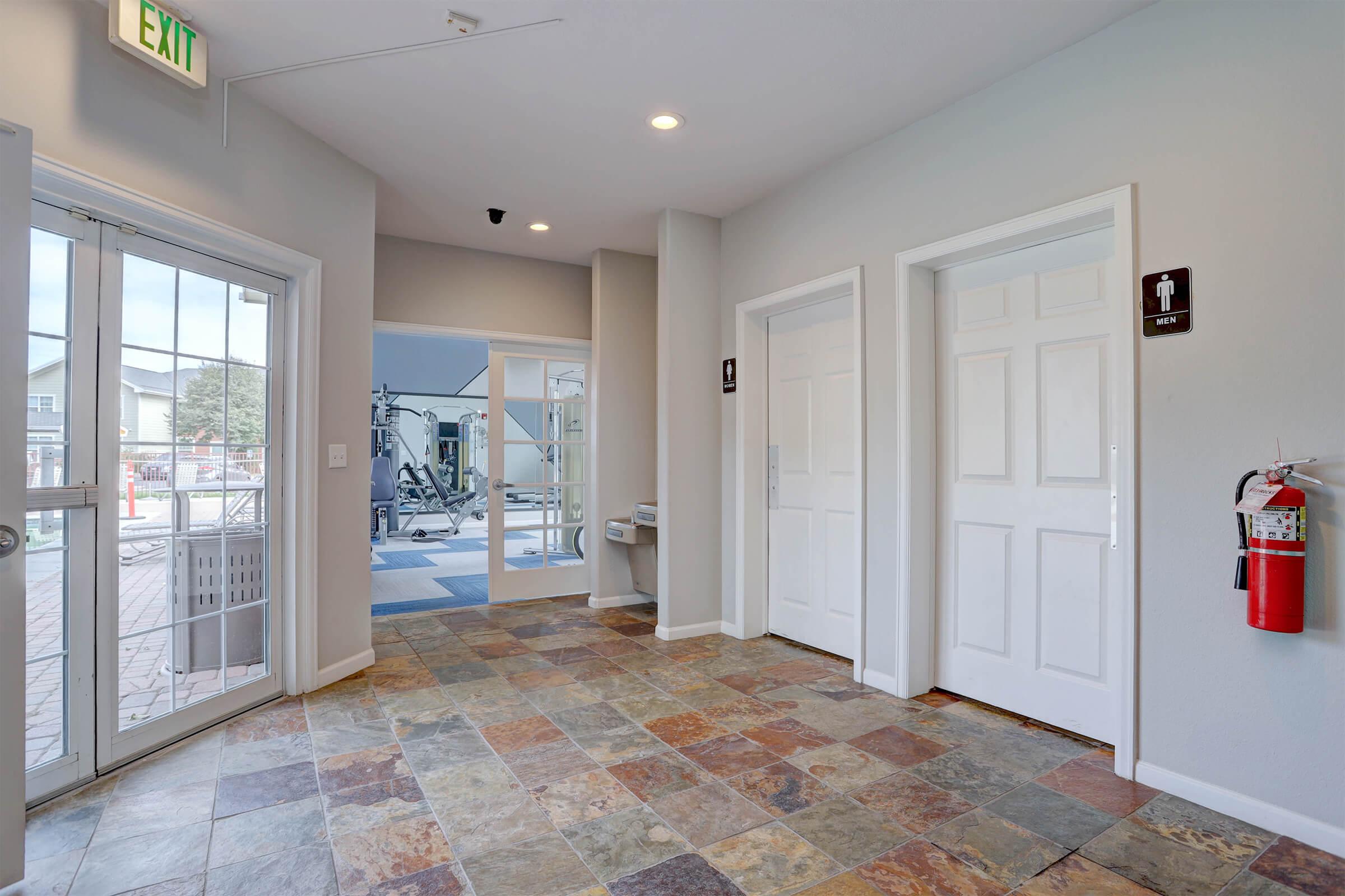 Interior view of a hallway featuring two closed doors labeled for restrooms, a glass entrance with exit sign, a fire extinguisher mounted on the wall, and a tiled floor. In the background, a gym area can be seen through a glass door.