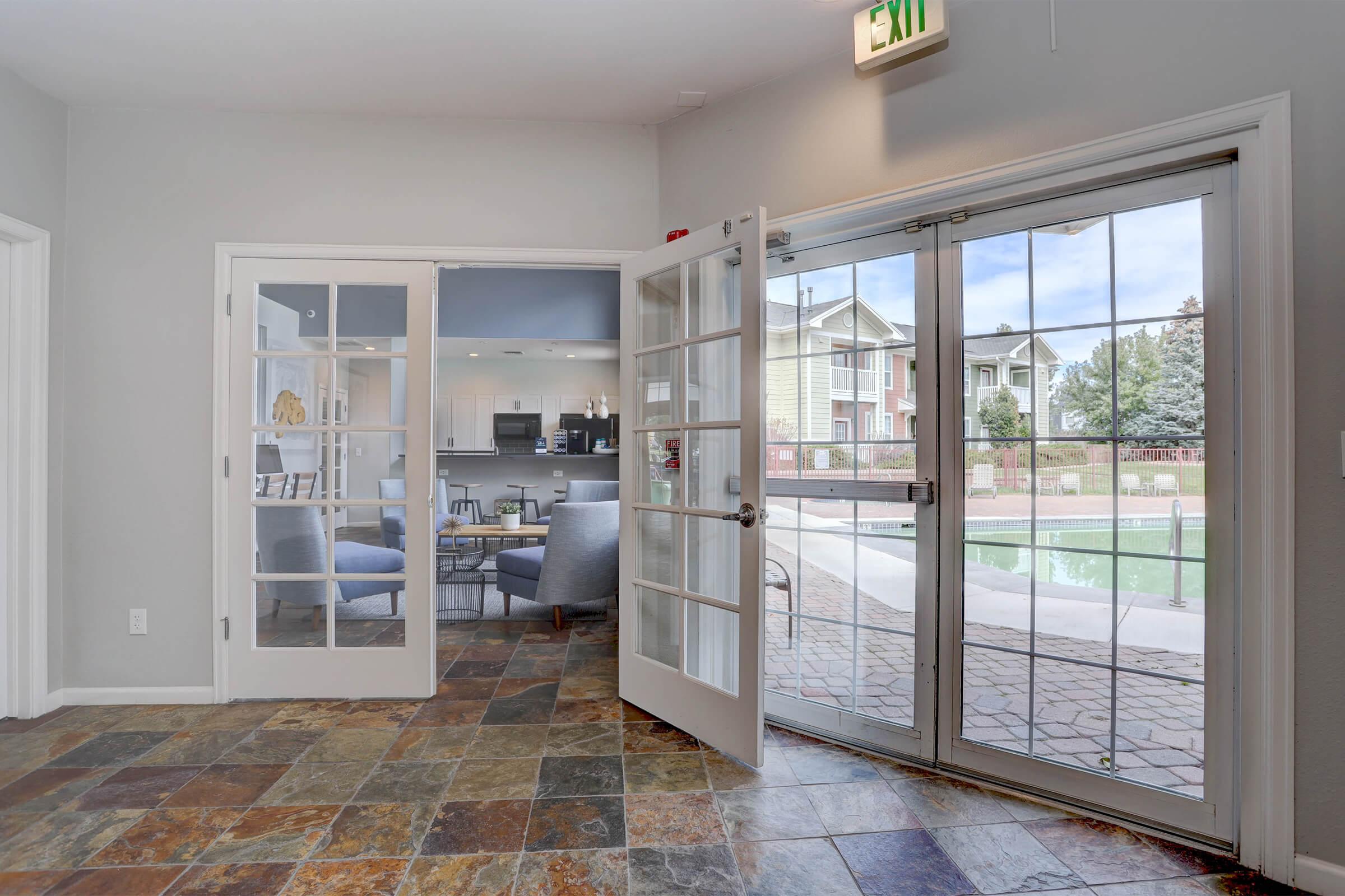A spacious interior view from a room with French doors opening to an outdoor area. The floor features slate tiles, and through the doors, a pool and a landscaped area are visible. Soft natural light fills the room, creating a welcoming atmosphere.
