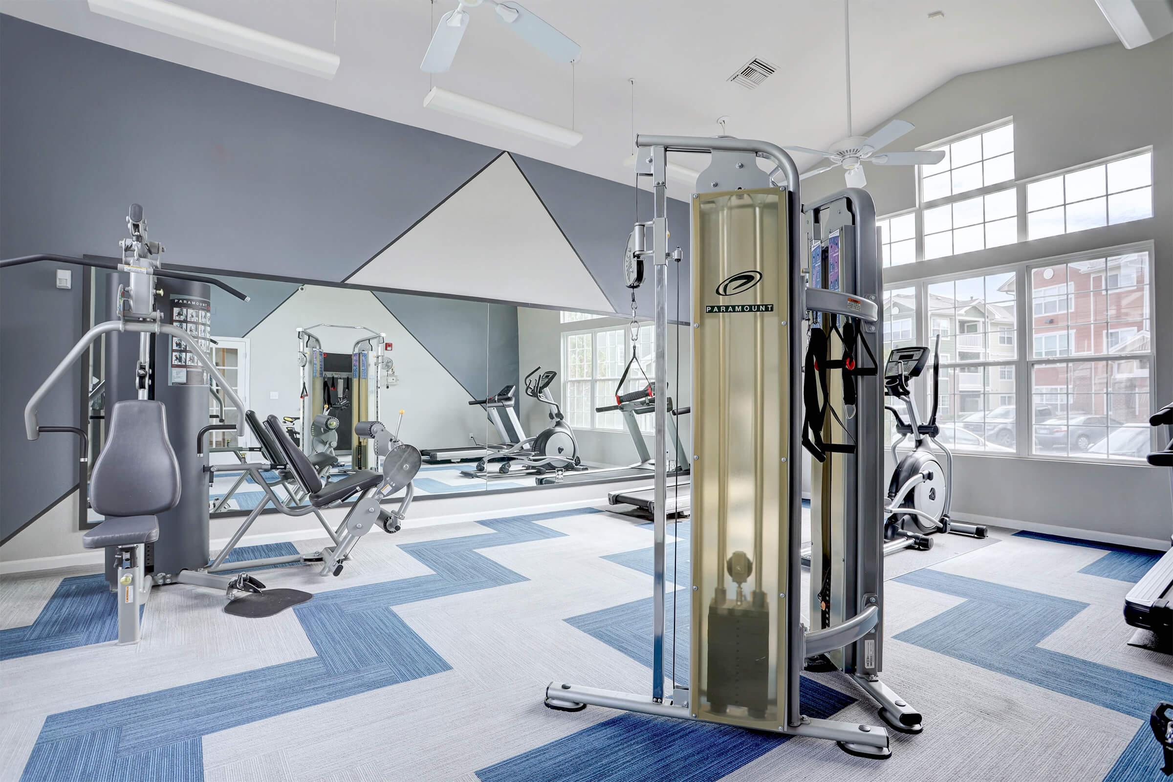 A modern gym interior featuring various fitness equipment, including weight machines, cardio machines, and a mirror. The space has a bright and airy feel, with large windows allowing natural light and a blue and gray color scheme on the flooring. Ceiling fans provide ventilation.