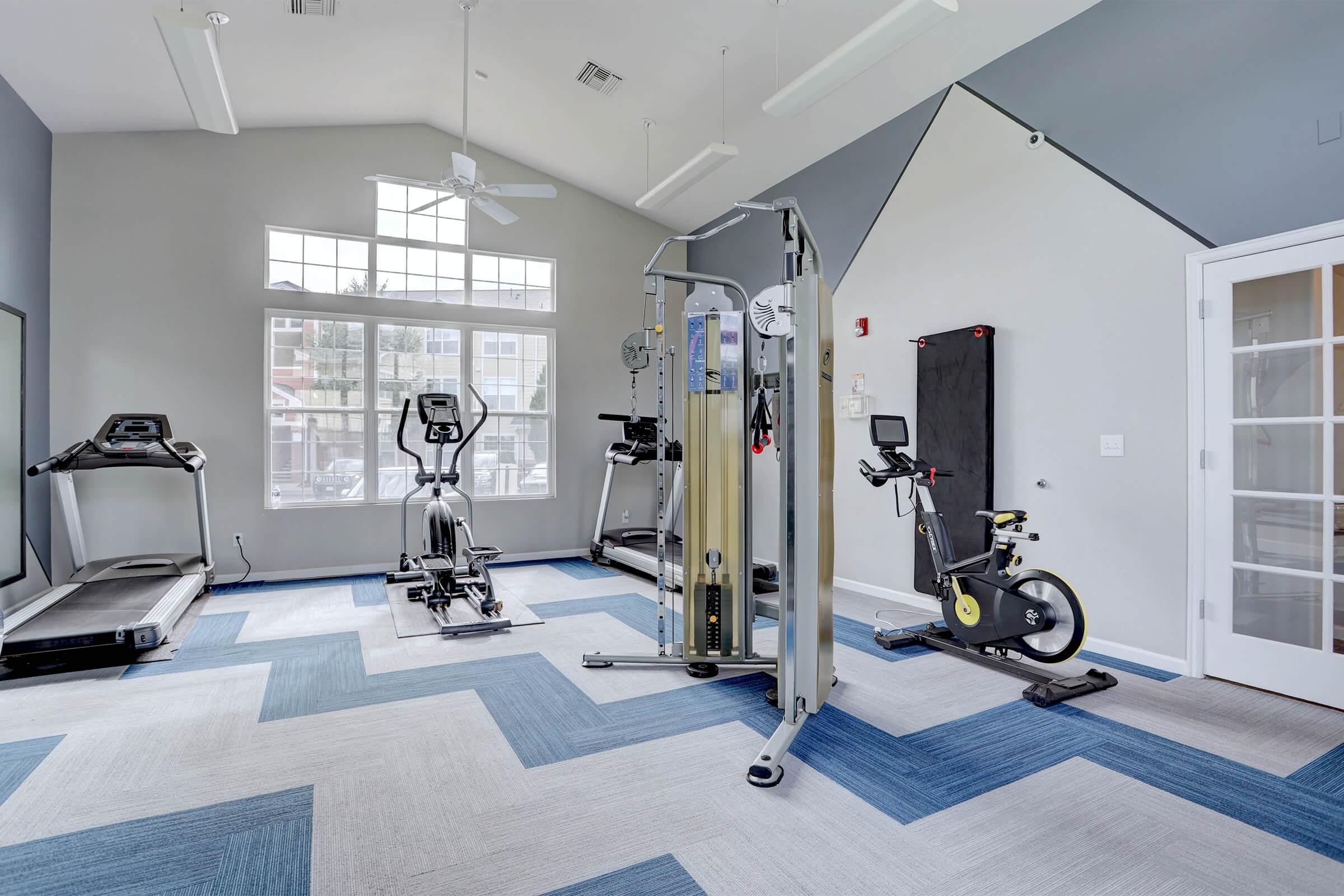 A well-lit gym interior featuring various exercise equipment, including treadmills, an elliptical machine, a strength training machine, and a stationary bike. The floor has a modern blue and gray patterned design, and large windows allow natural light to fill the space.