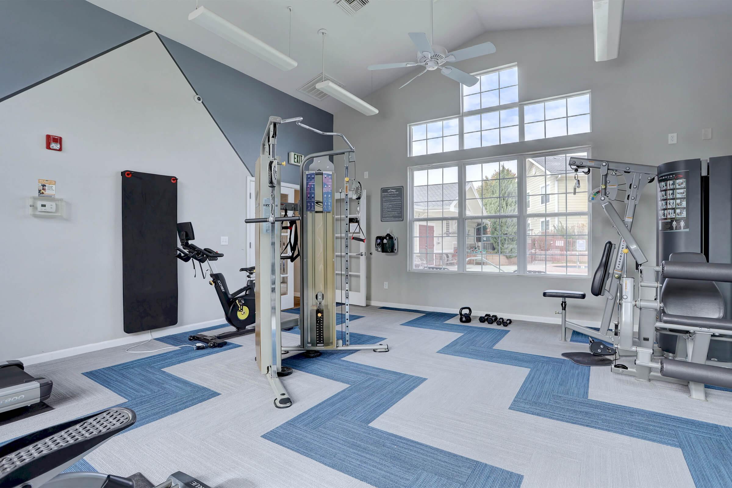 A modern gym interior featuring various fitness equipment, including a multi-station weight machine, a stationary bike, and free weights. The space has large windows allowing natural light, with a mix of blue and gray flooring patterns to enhance the aesthetic.