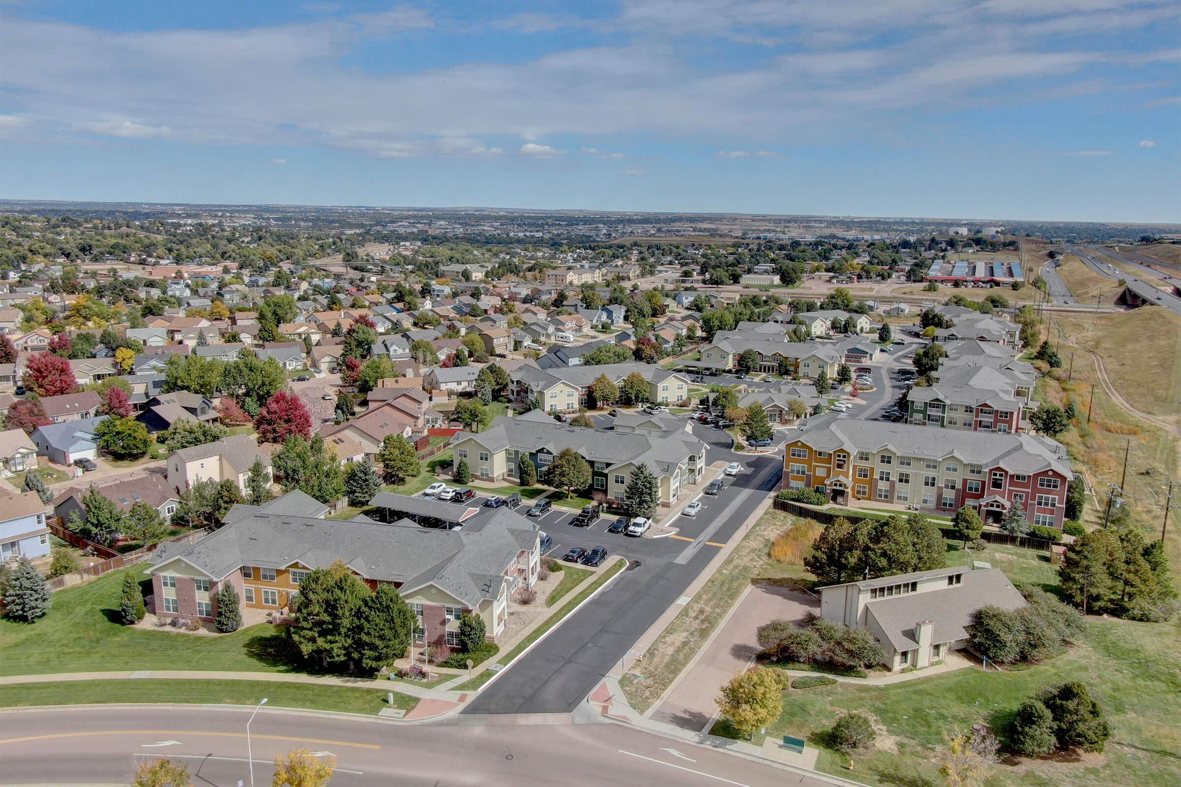 Aerial view of a suburban neighborhood featuring various residential buildings, including apartments and houses. The landscape includes trees in autumn colors and well-maintained streets. A highway is visible in the distance, with open fields beyond the neighborhood. The sky is clear with a few scattered clouds.