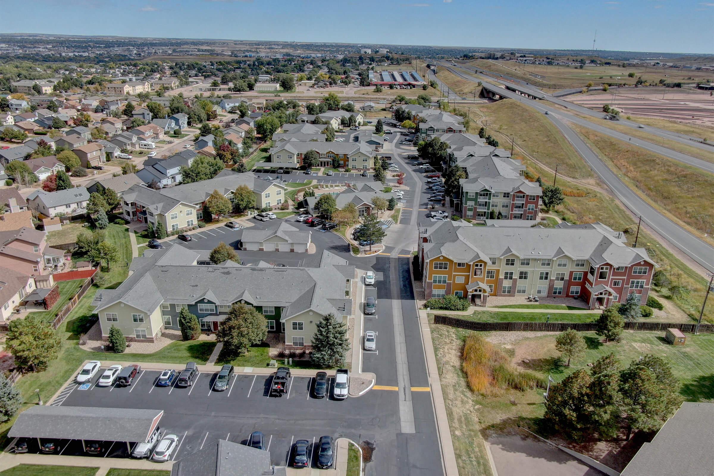 Aerial view of a residential neighborhood featuring multi-story apartment buildings with various architectural styles and colors. The area includes parking lots, landscaped greenery, and nearby roadways. In the background, there are additional homes and open land, suggesting a suburban environment.
