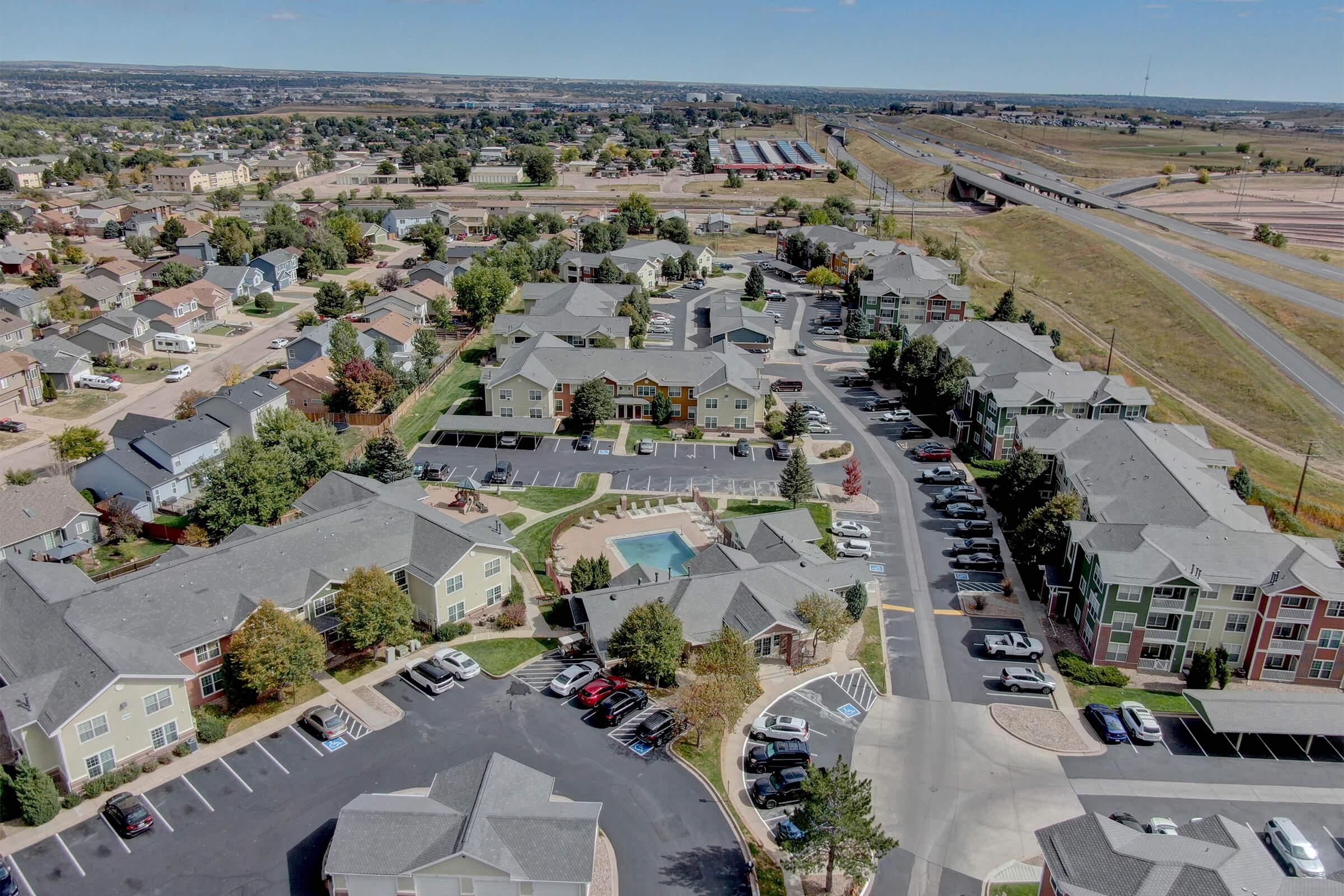 Aerial view of a residential neighborhood featuring multiple apartment buildings, parking lots, and a swimming pool. The area is surrounded by greenery, with a highway and open land visible in the background. The scene shows clear skies and a suburban layout, indicating a well-developed community.