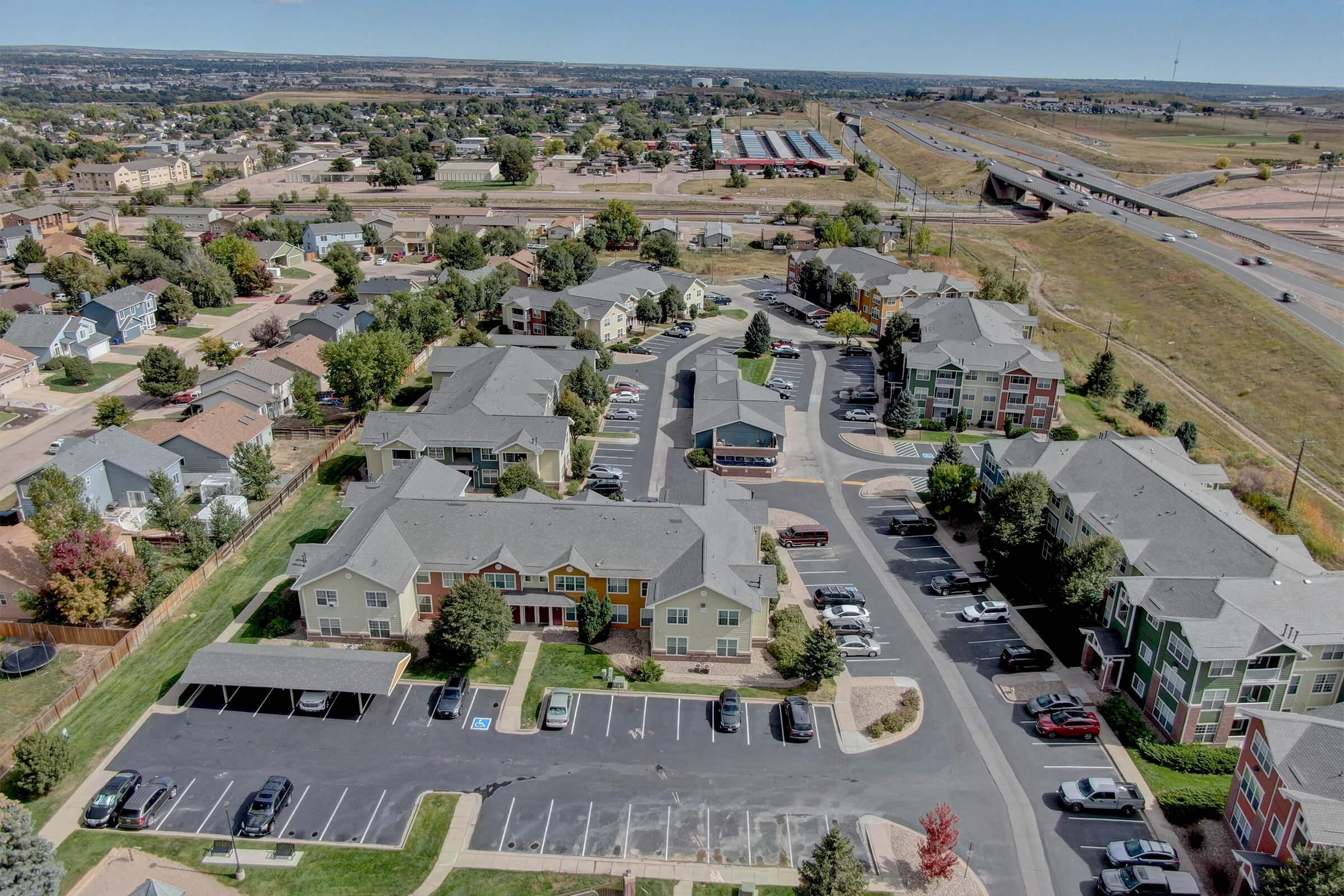 Aerial view of a suburban apartment complex with several multi-story buildings surrounded by parking lots and trees. In the background, there is a highway and residential areas, along with open land and some commercial buildings. The scene captures a clear day with blue skies.