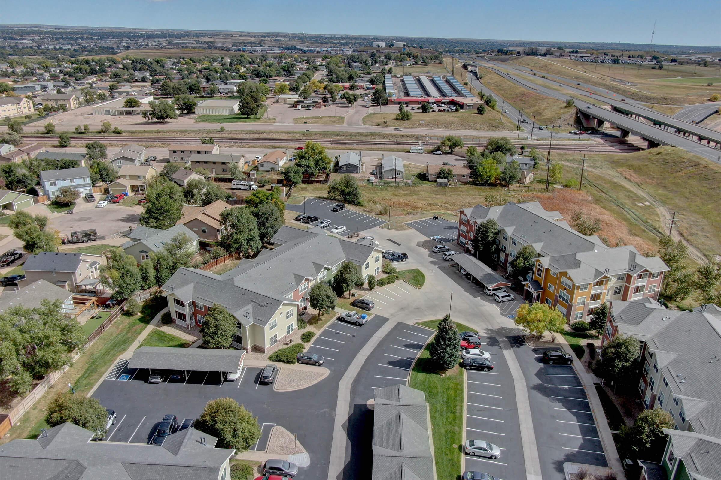 Aerial view of a residential area featuring several multi-story apartment buildings, parking lots, and surrounding greenery. In the background, there are commercial buildings, a highway, and open land, indicating a mix of urban and suburban development. Clear blue sky above.