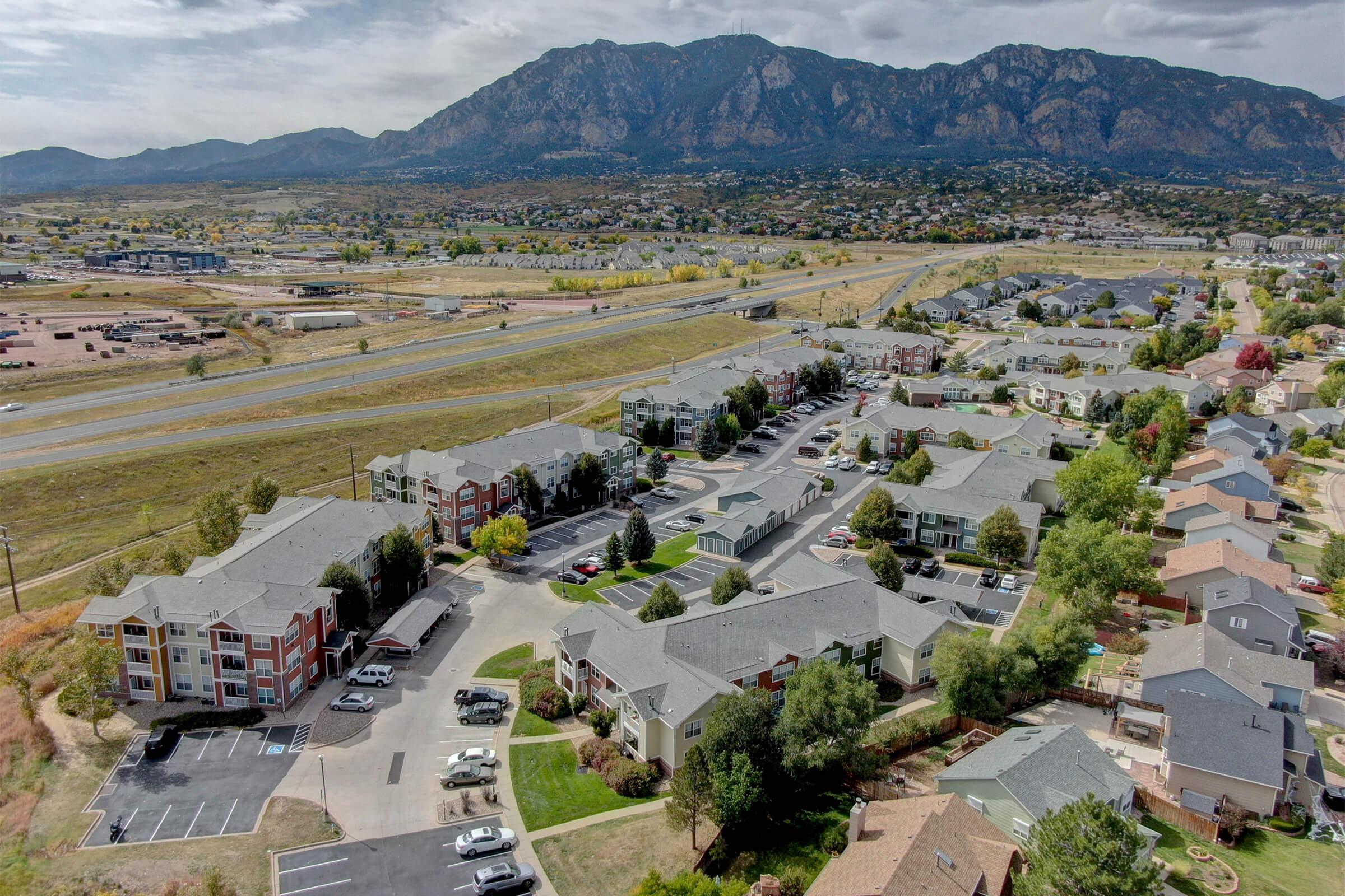 Aerial view of a suburban neighborhood featuring various low-rise apartment buildings and well-maintained landscaping. In the background, there are mountains and a clear sky, while in the foreground, parking lots and vehicles are visible, along with open green spaces and roads.